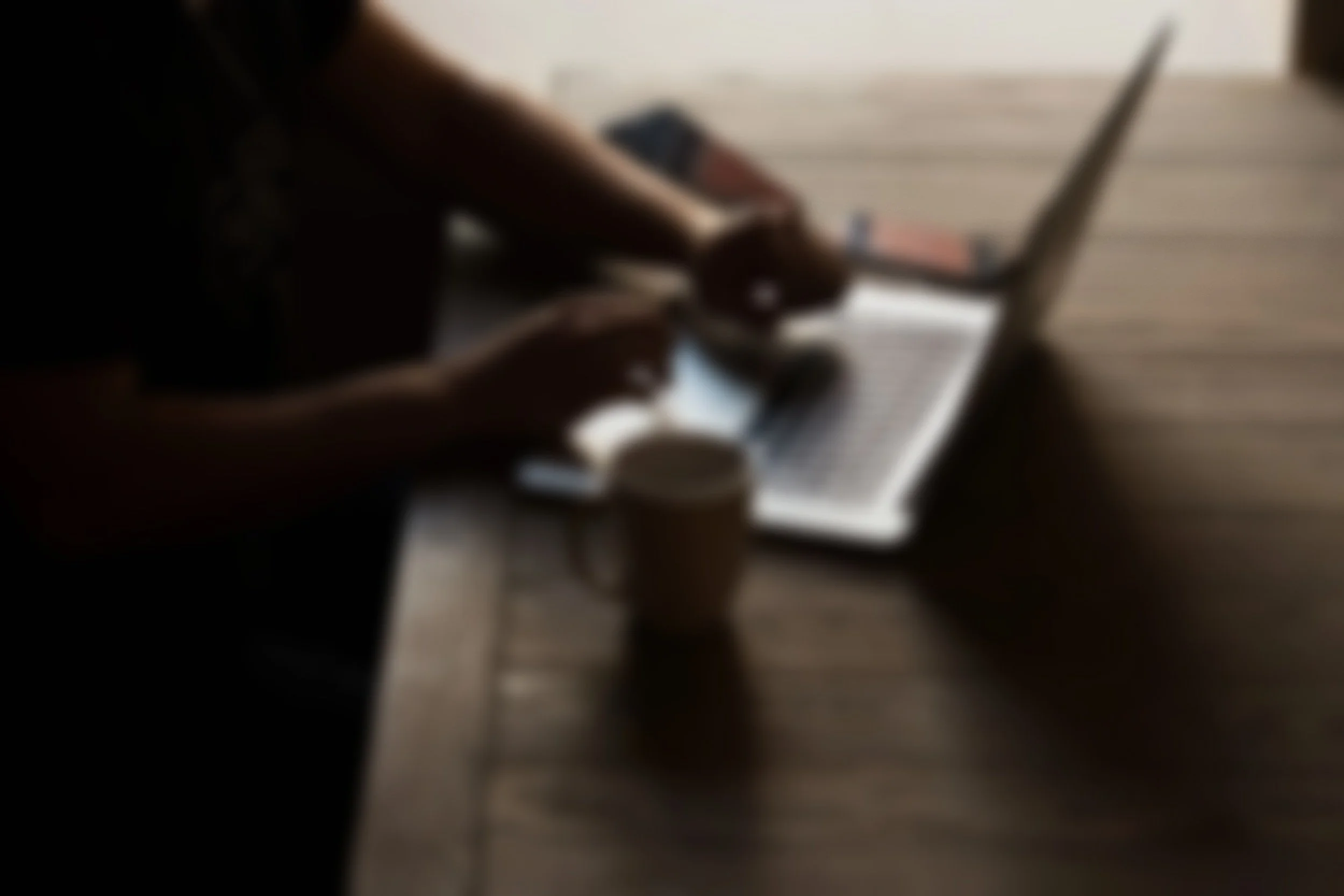 Person som använder en bärbar dator på ett träbord, med två mobiltelefoner och en kaffekopp.