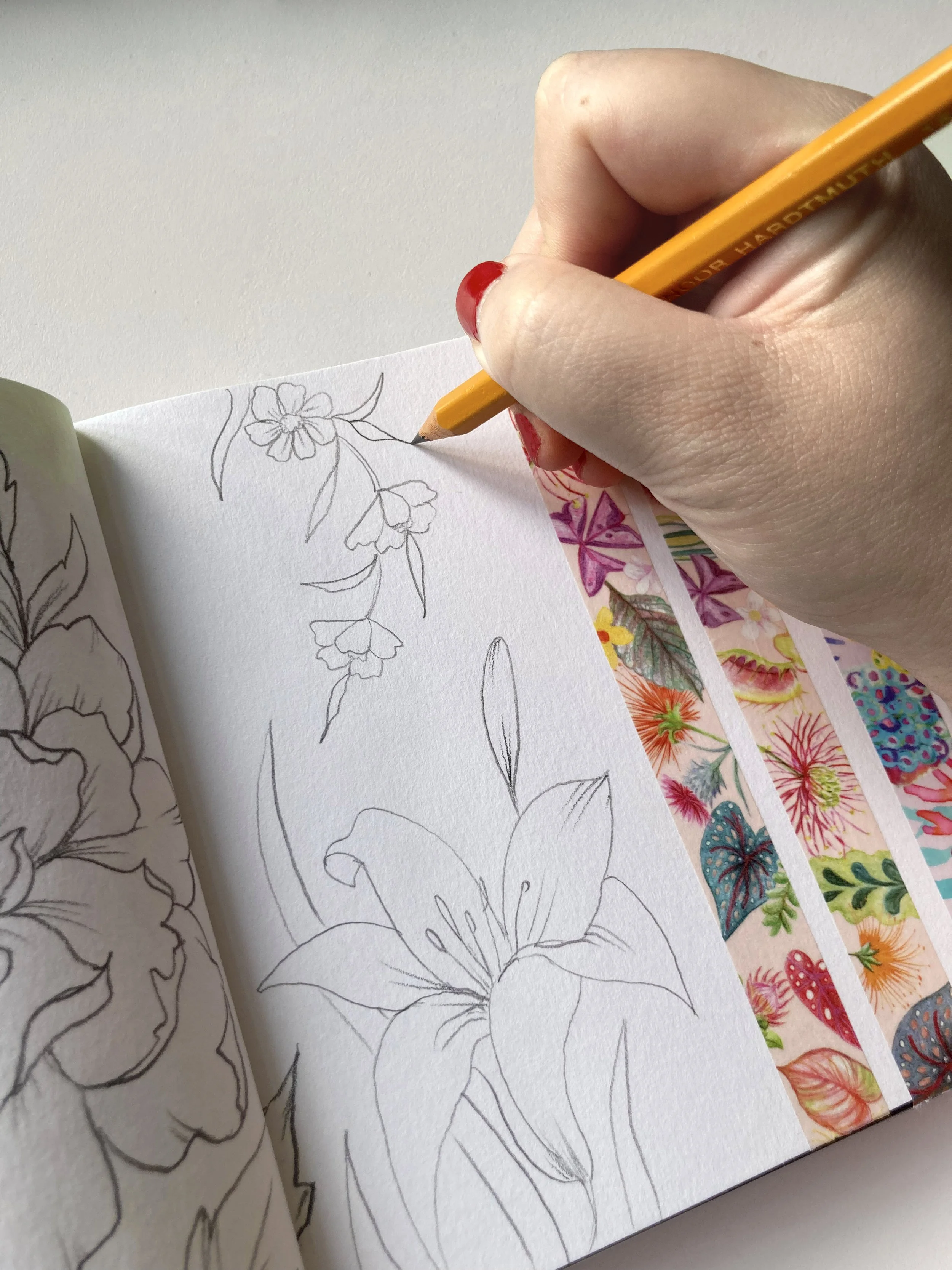 A hand with red painted nails using a yellow pencil to trace a flower outline in a sketchbook. The sketchbook is open to a page with floral sketches and colorful floral stickers on the right side.