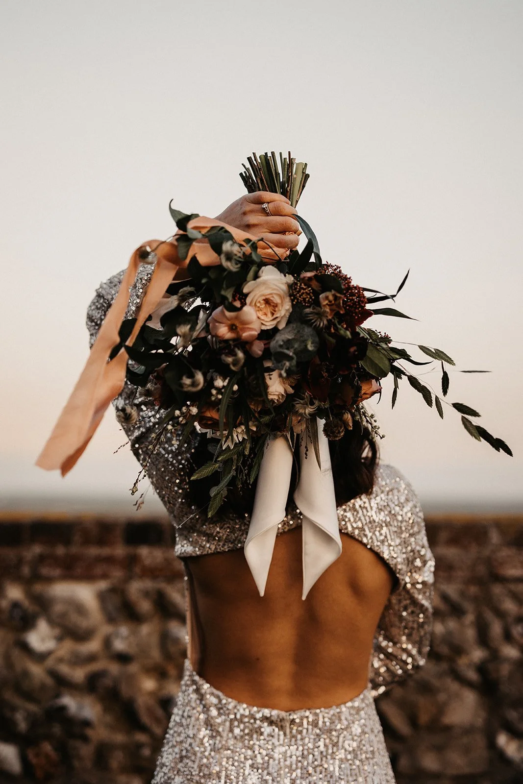 Editorial dark moody shot of bride facing away from camera holding floral bouquet over shoulder
