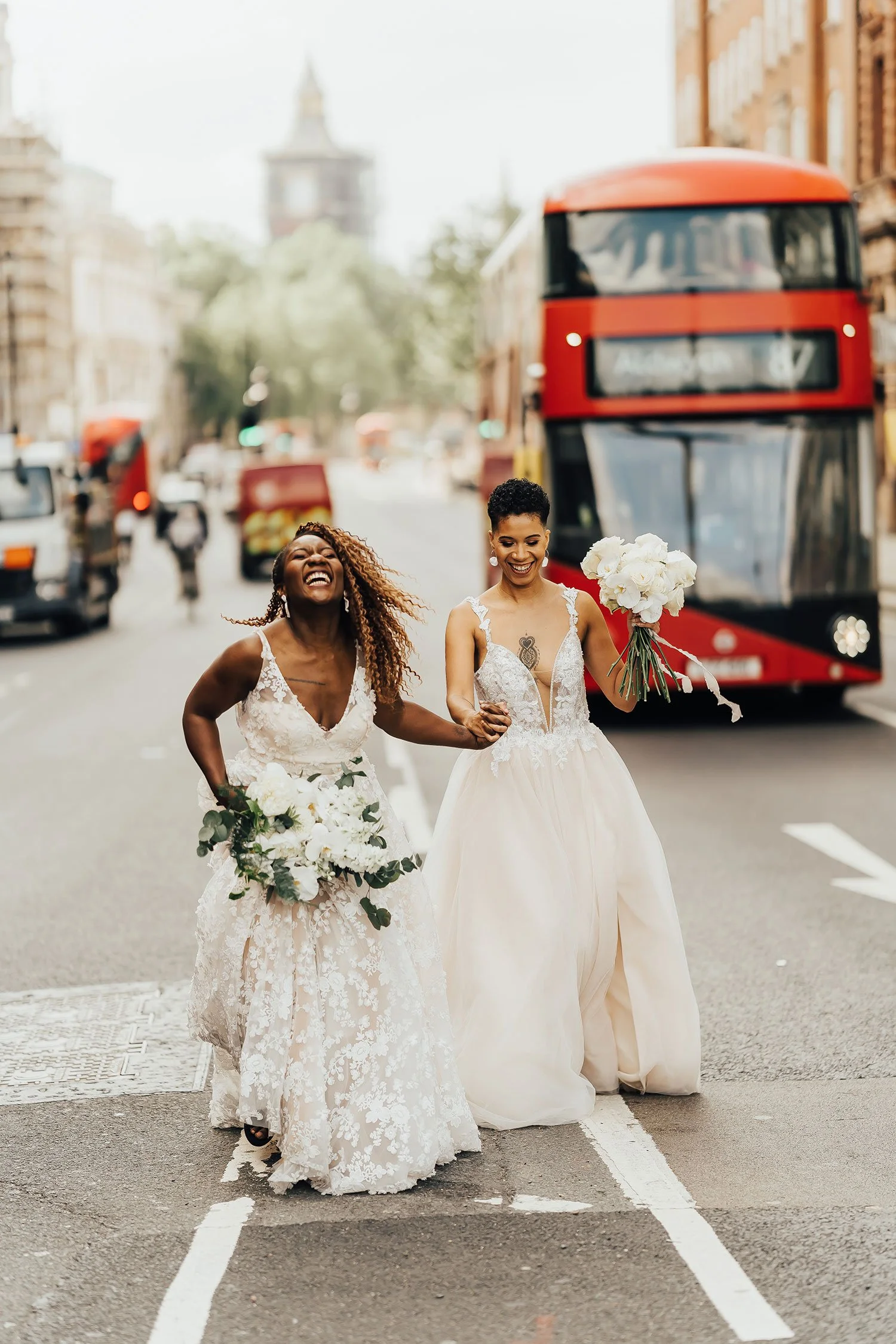 Two brides holding hands laughing with floral bouquets and London bus
