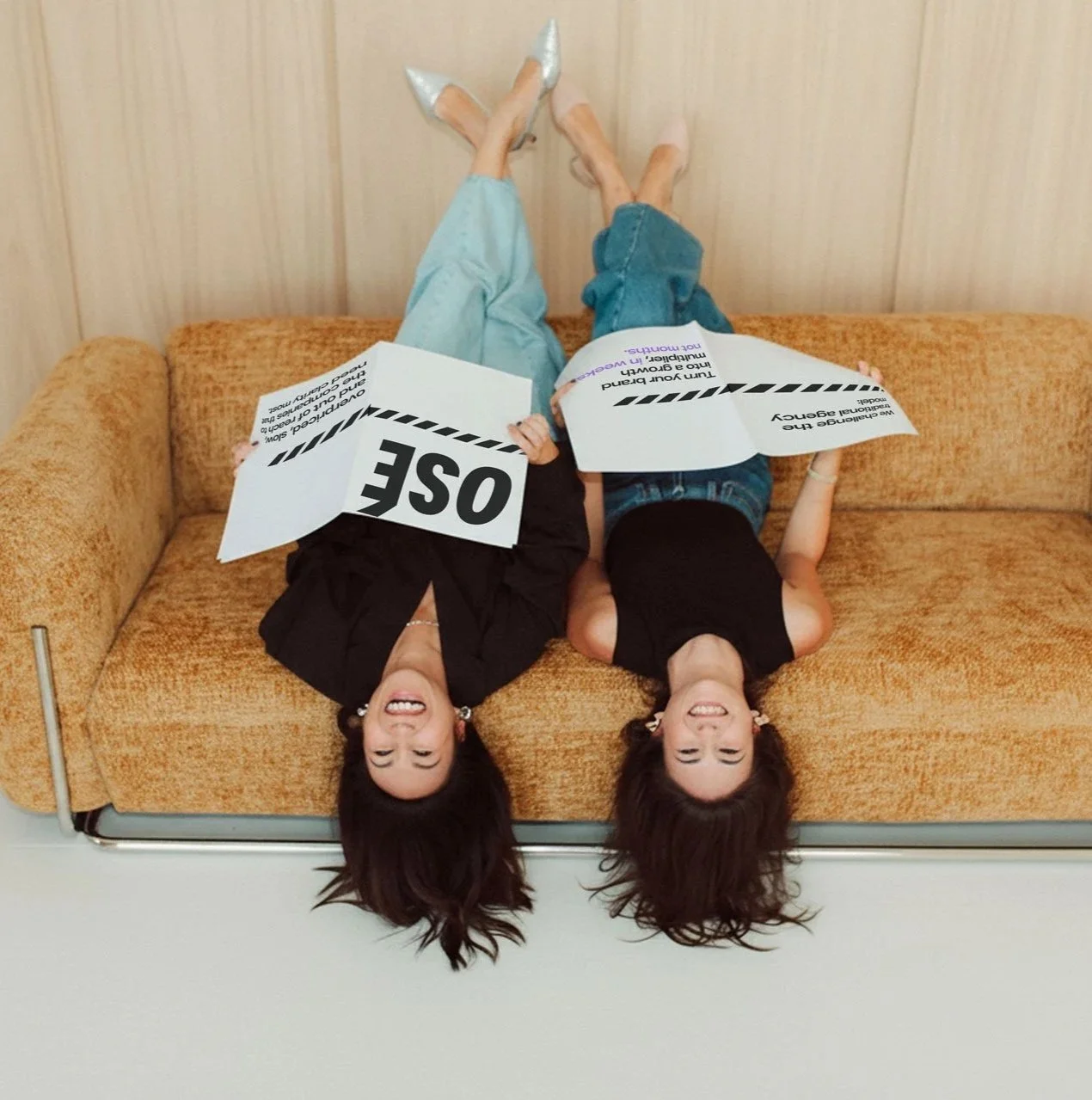 Two women lying upside down on a vintage orange sofa, holding protest signs with one reading 'OBE' and the other 'We are standing here in support of your campaign'.