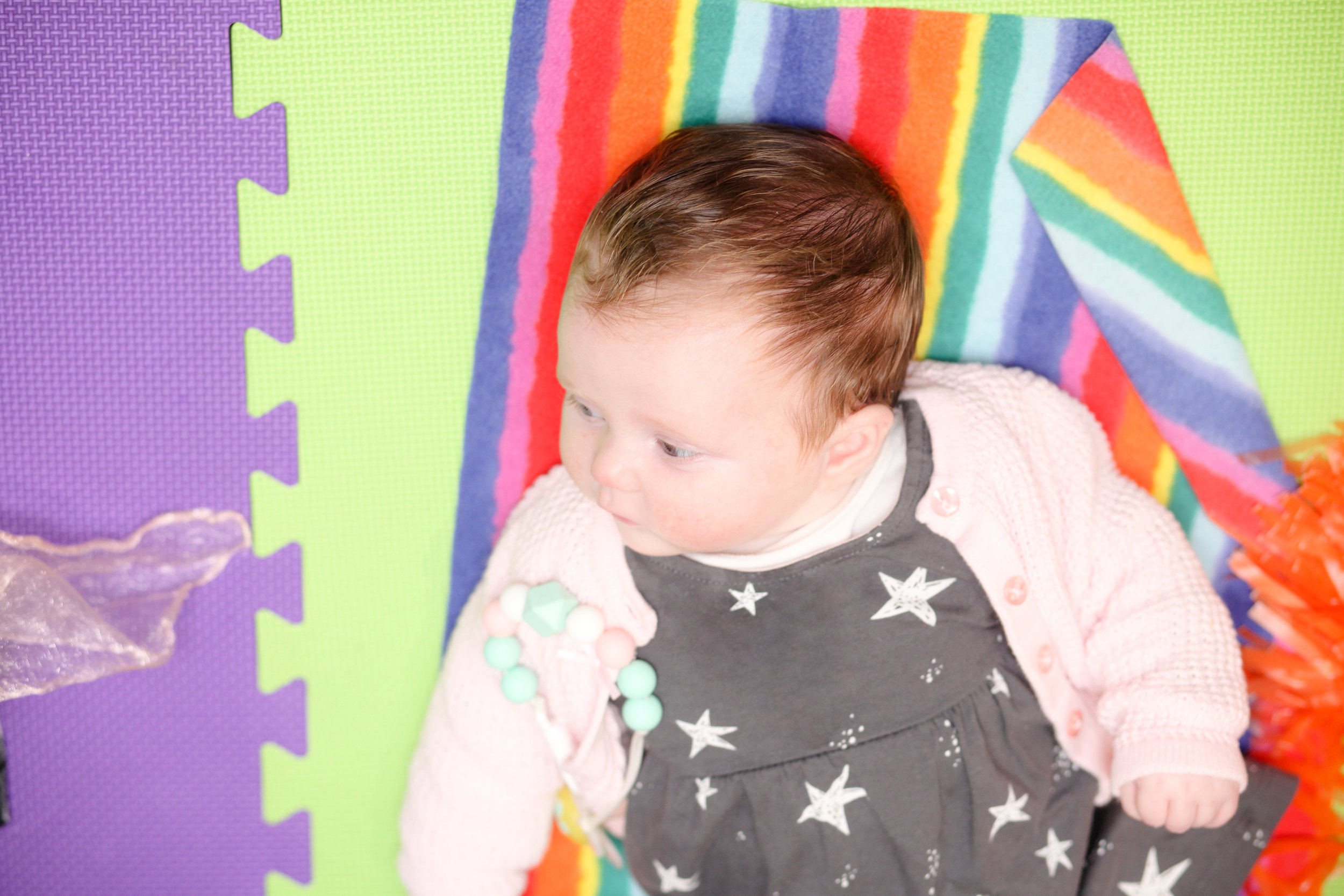 A baby girl with reddish-brown hair lying on a rainbow-colored striped blanket on a soft foam play mat.