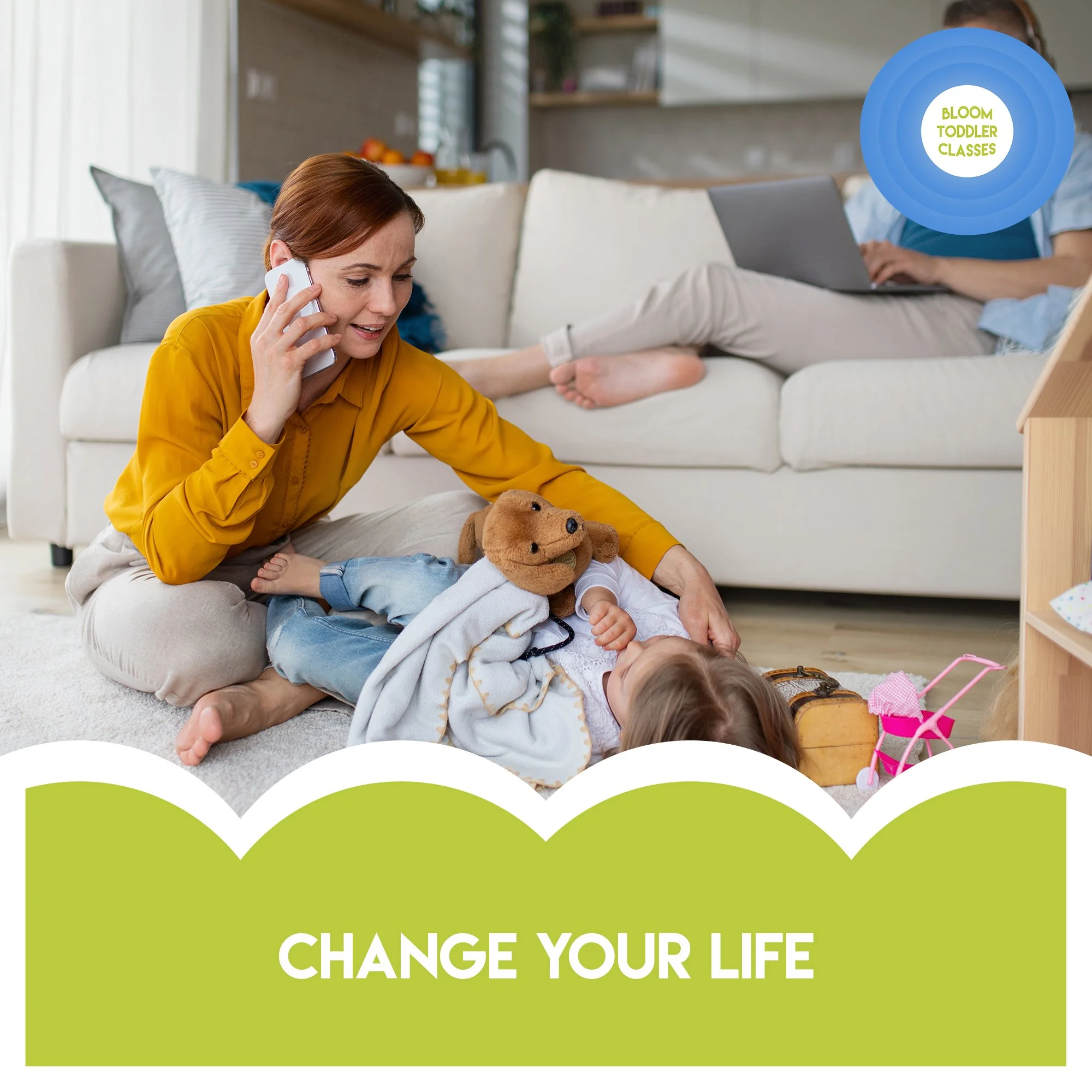 A woman sitting on the floor with a young child, both playing with toys in a living room. The woman is on her knees, talking on the phone, smiling. The child is lying down, dressed in light-colored clothes, with a stuffed puppy toy nearby. In the background, another person is sitting on the couch with a laptop. The room has a cozy, homey feel with cushions, shelves, and a bowl of fruit.