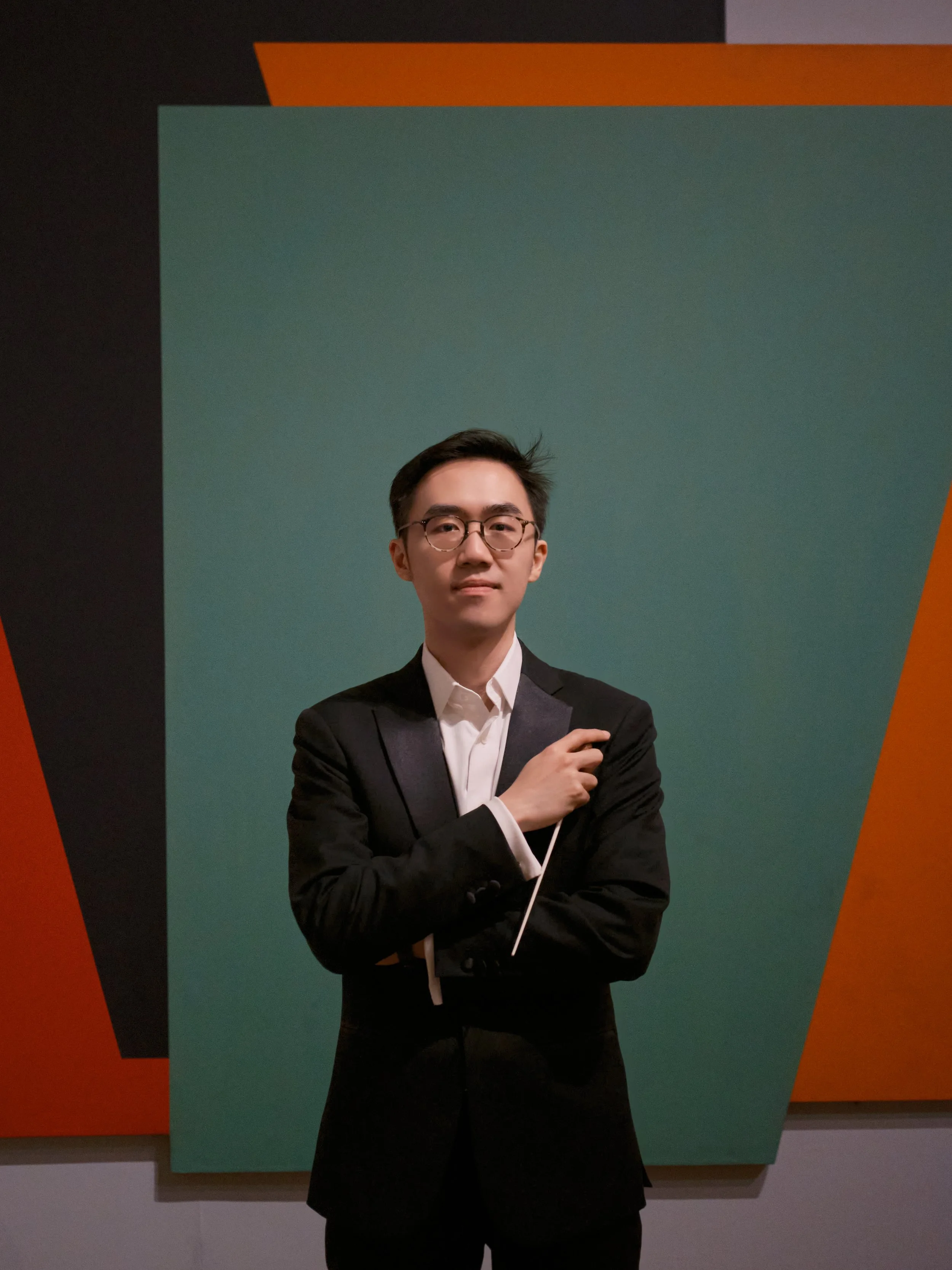 A young man in a black suit and glasses standing in front of colorful geometric wall art.