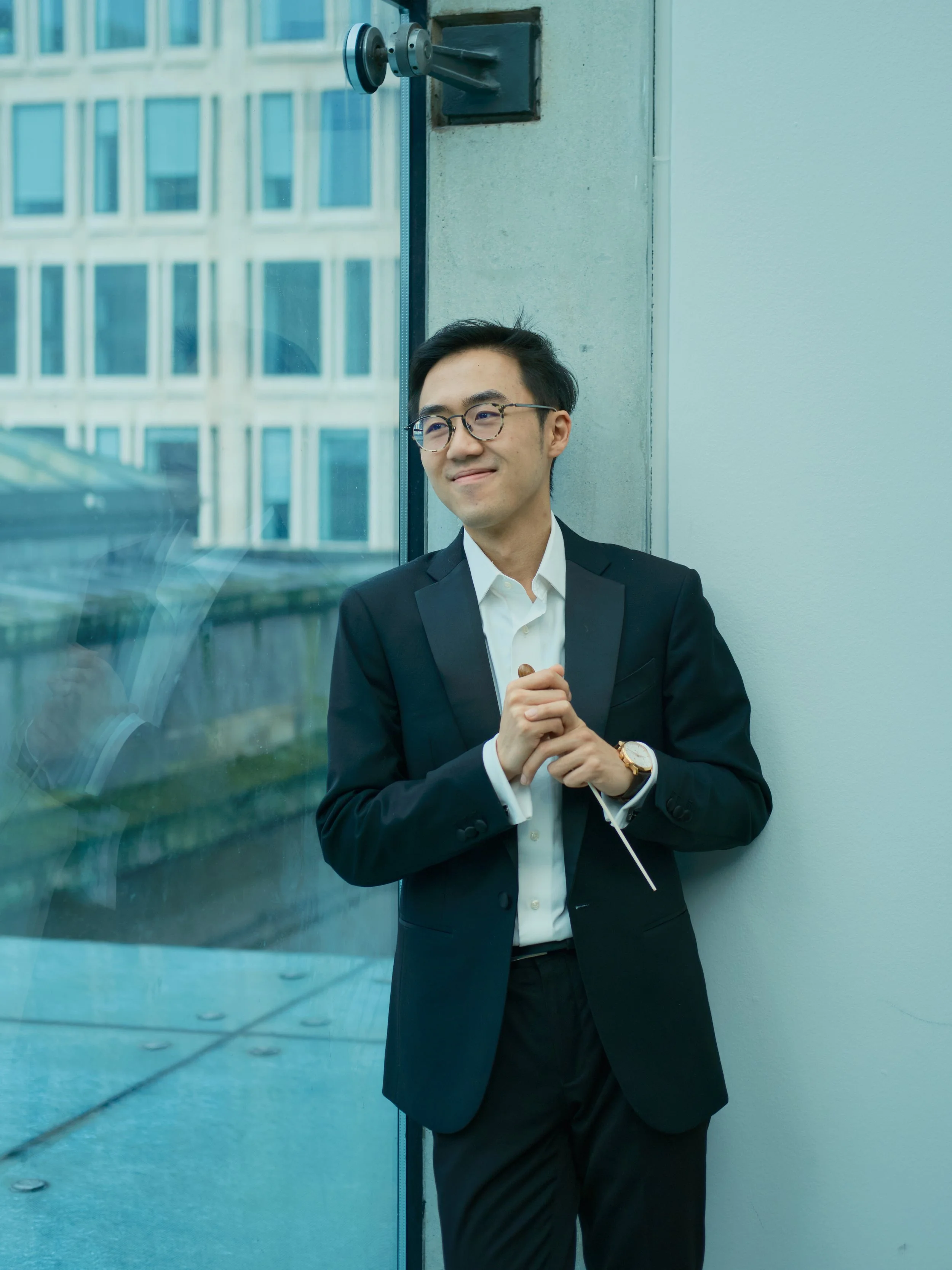 A young man in a black suit and white shirt stands by a glass wall, smiling softly and looking to the side.