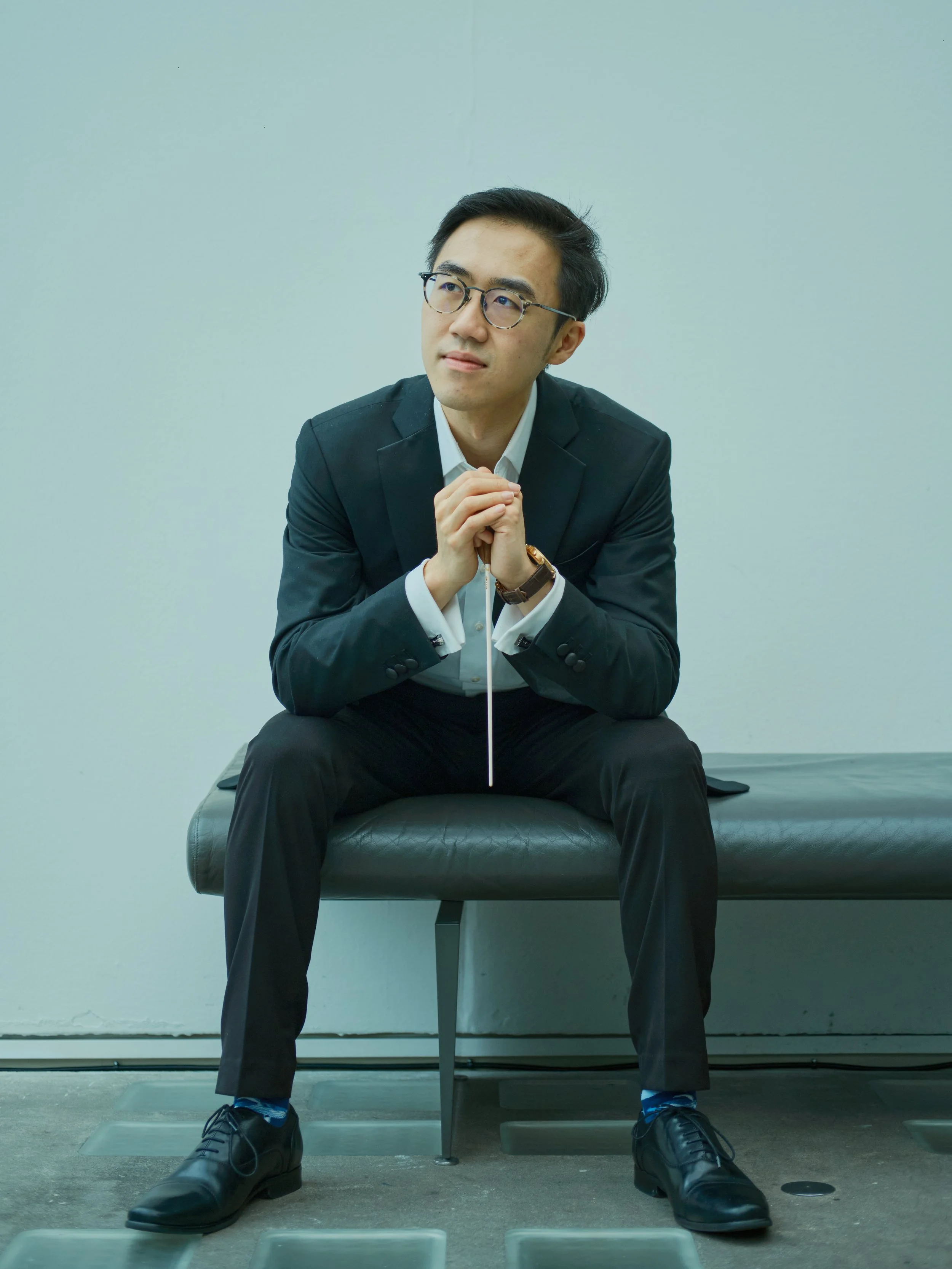 A man dressed in a black suit and white shirt, sitting on a black bench in a room with plain white walls, with his hands clasped together while holding a baton and looking thoughtfully to the side.