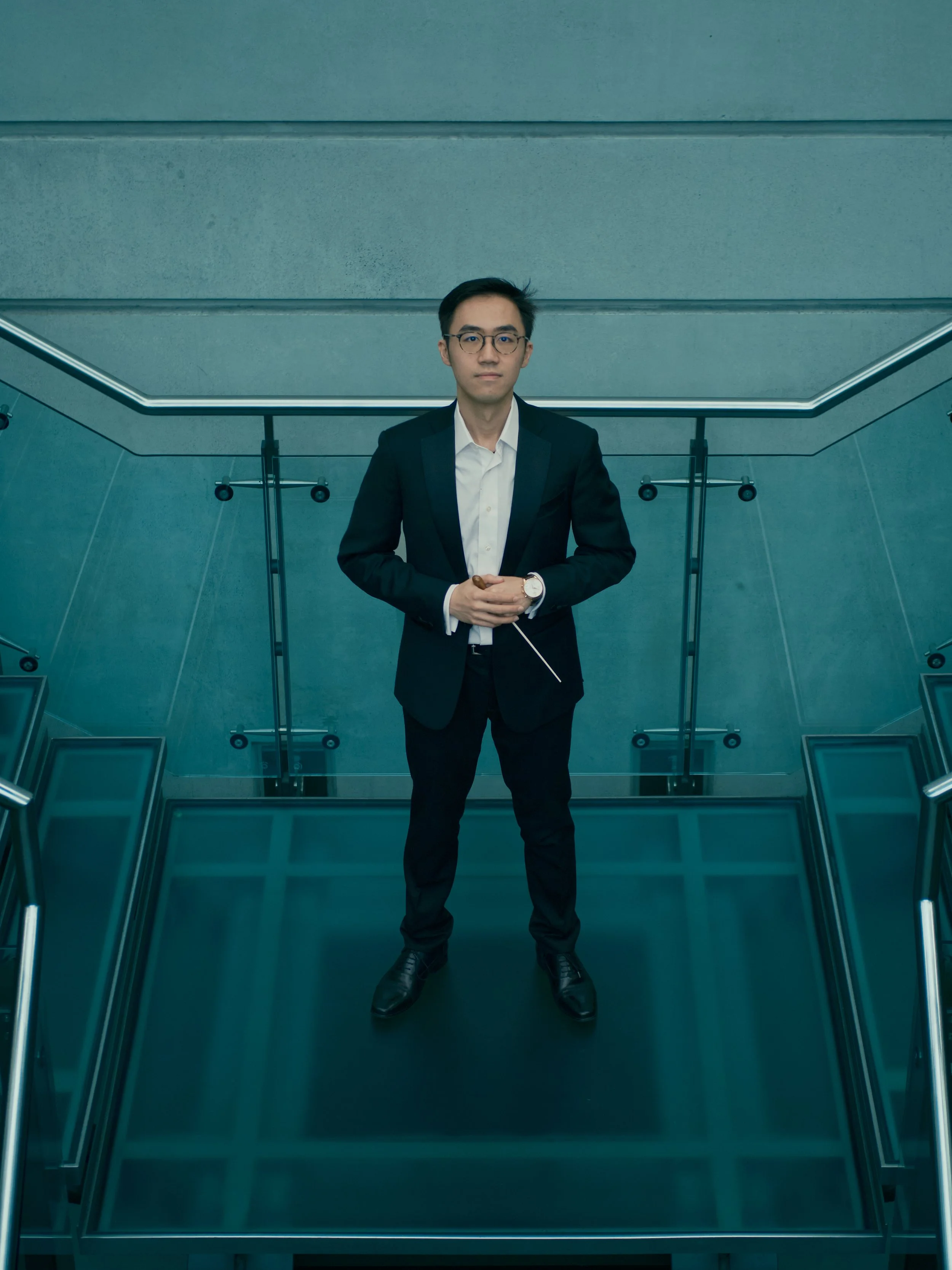 A man in a black suit and white dress shirt stands on a glass staircase in a modern, minimalist building. He is holding a baton in his right hand and wearing glasses and a watch.