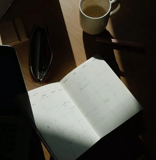 Open monthly planner on a wooden desk with a cup of coffee, a black pencil case, and a closed laptop in sunlight.
