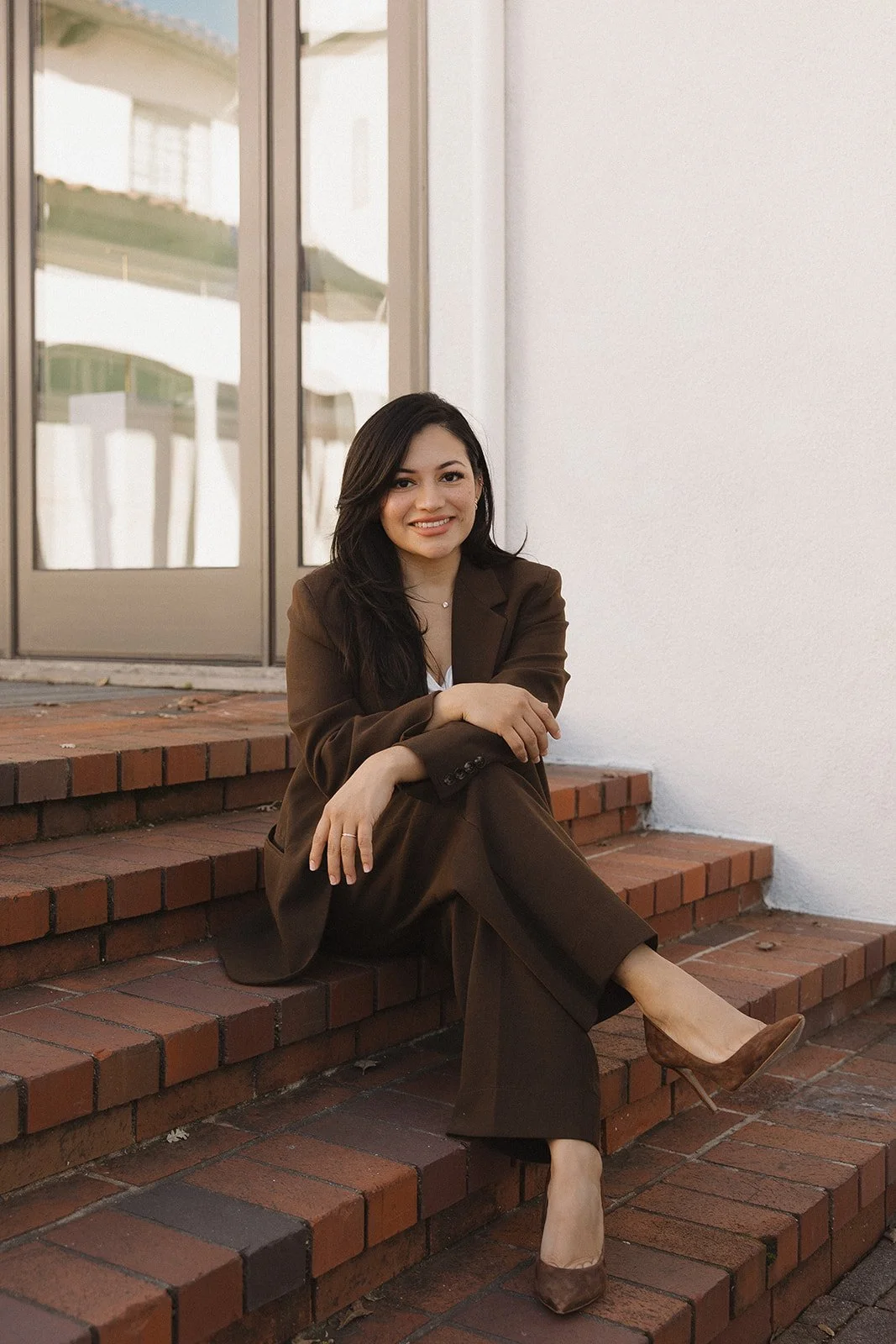 A woman in a brown suit sitting on brick steps outside a building, smiling at the camera.