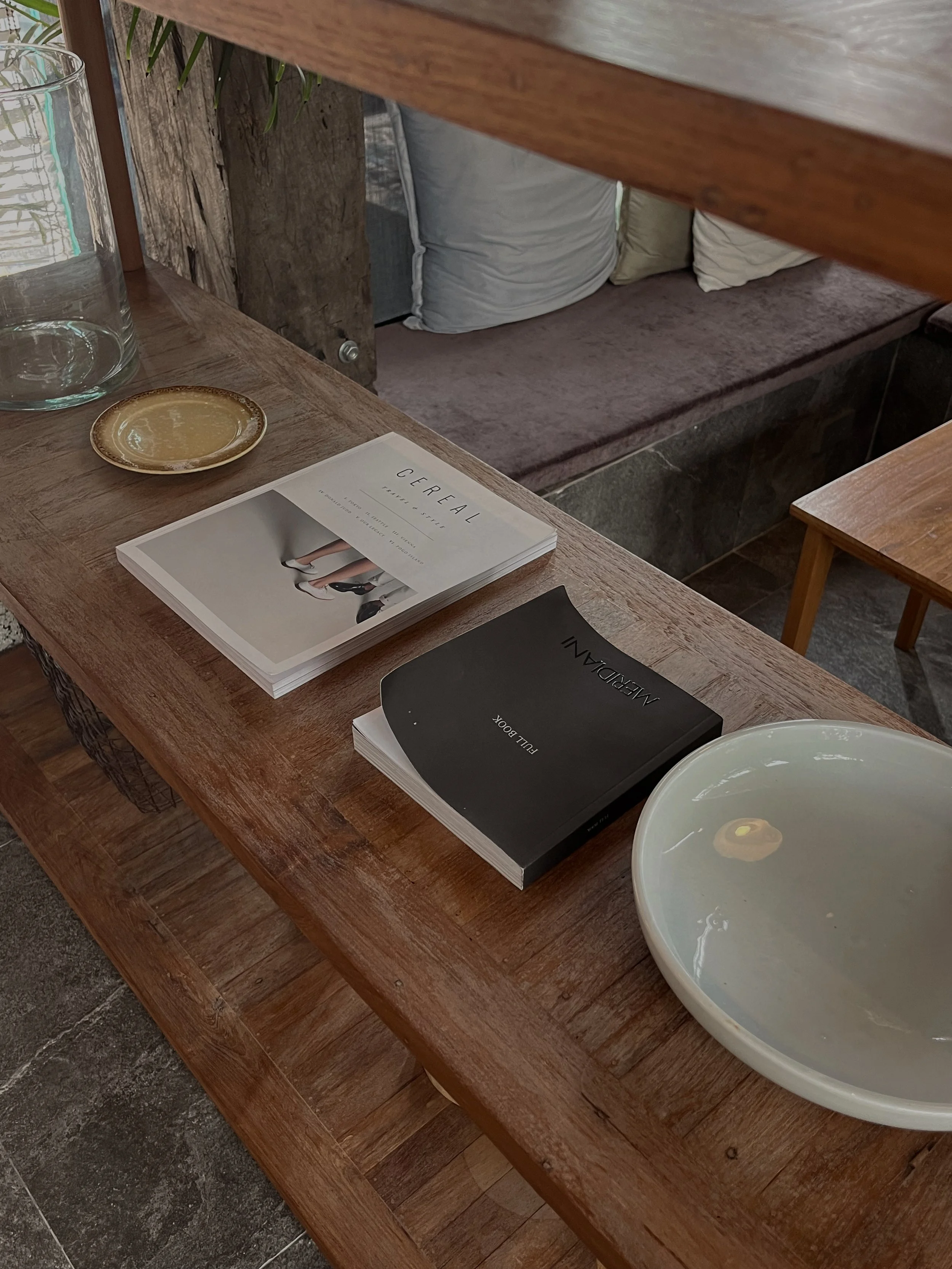A wooden table with magazines, a ceramic dish, a glass container, and a bowl with a small yellow object inside. In the background, there is a cushioned bench with pillows.