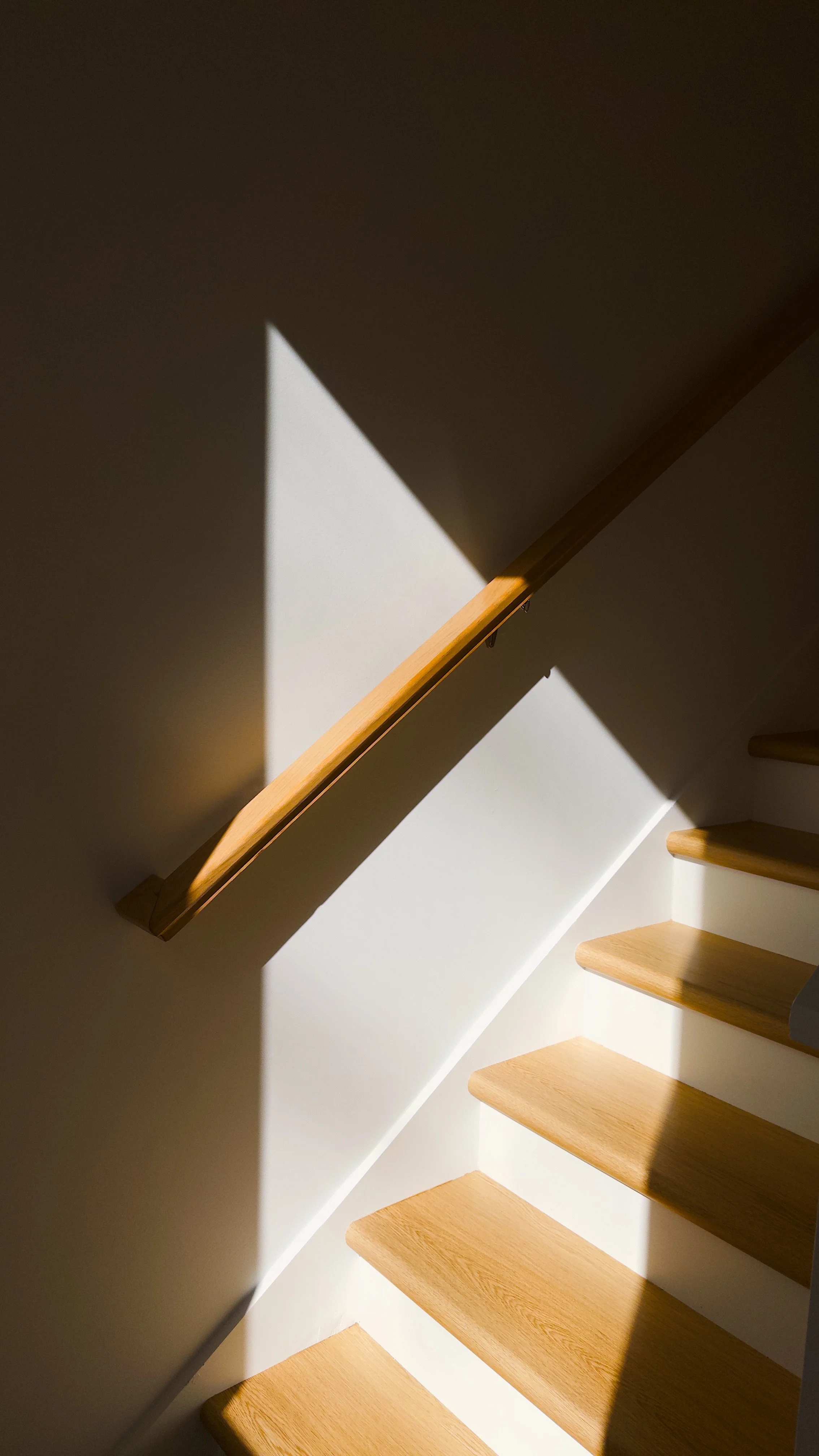 Sunlight casting diagonal shadows on wooden stairs and white wall, emphasizing geometric shapes.