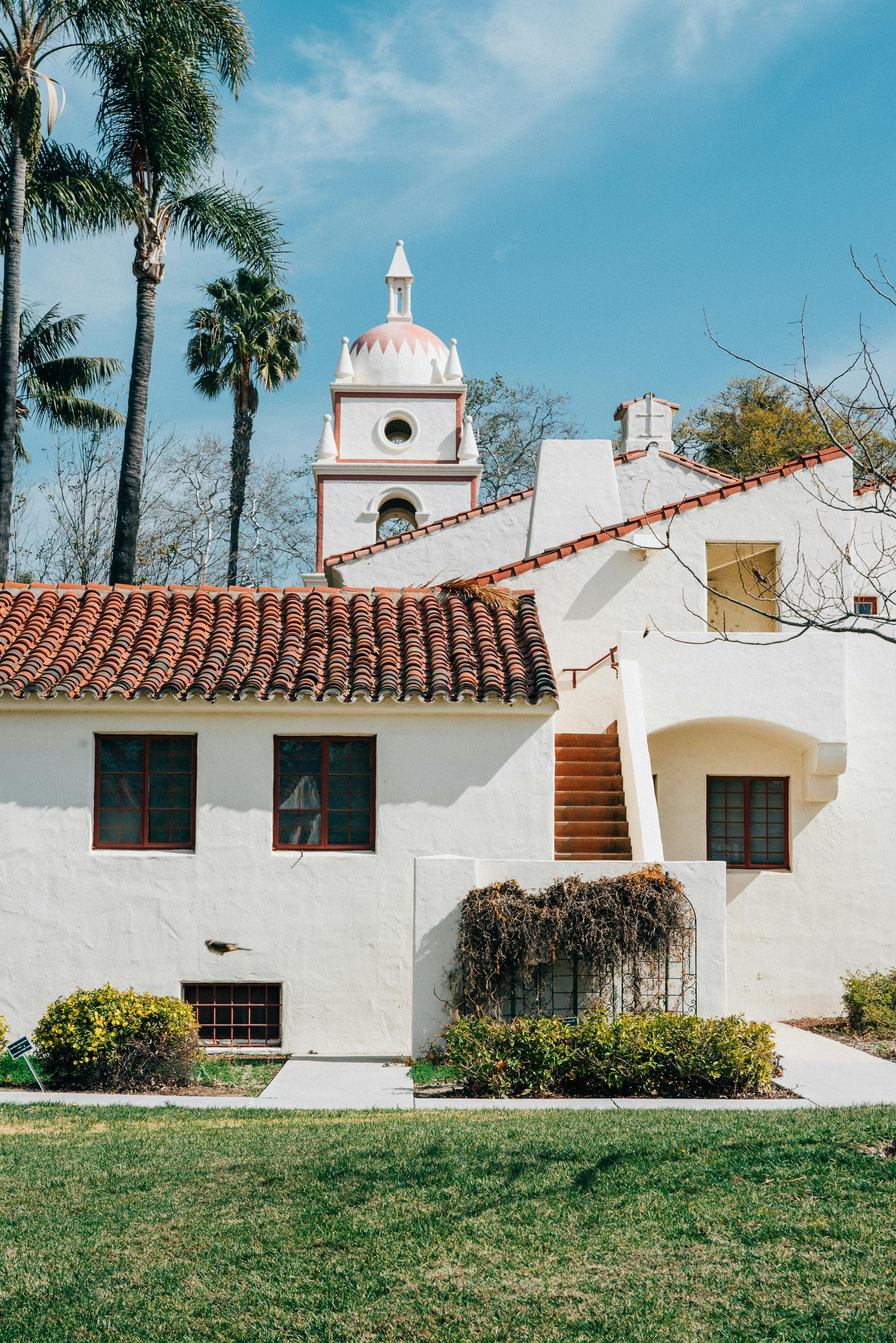 A white stucco building with red-tile roof, small windows, and a tower with a pink and white domed top, set against a blue sky and tall palm trees.