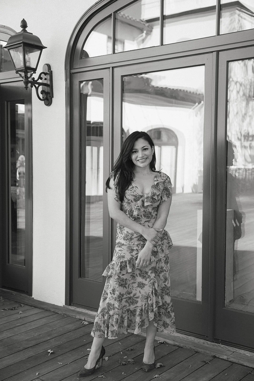 A woman standing outdoors in front of glass doors and window, wearing a floral dress and high heels, smiling at the camera.