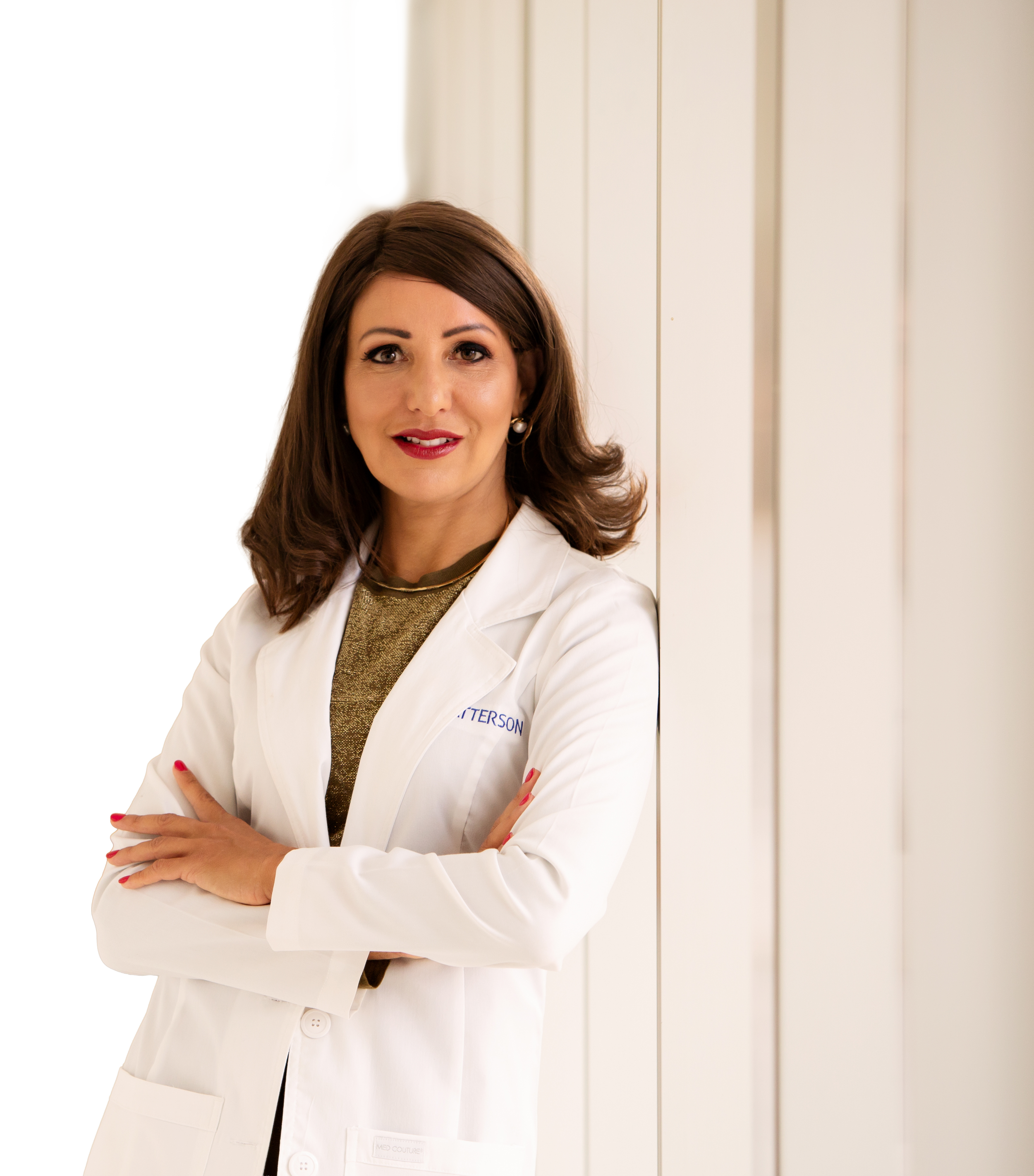 A woman with brown hair in a white lab coat standing with arms crossed, smiling, against a light background with vertical white panels.