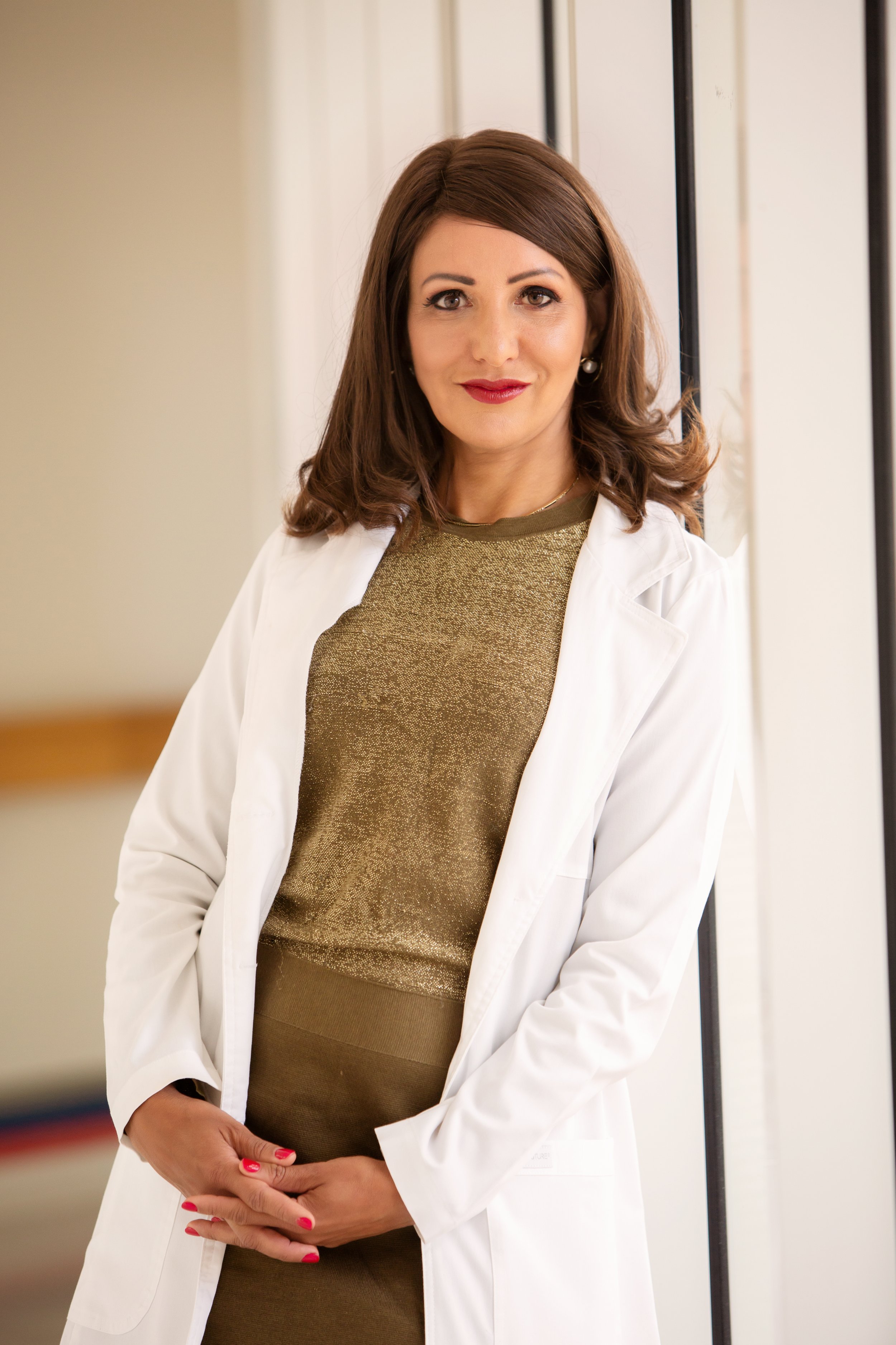 A woman with shoulder-length brown hair, wearing a white lab coat over a gold shirt, standing indoors near a railing and smiling at the camera.