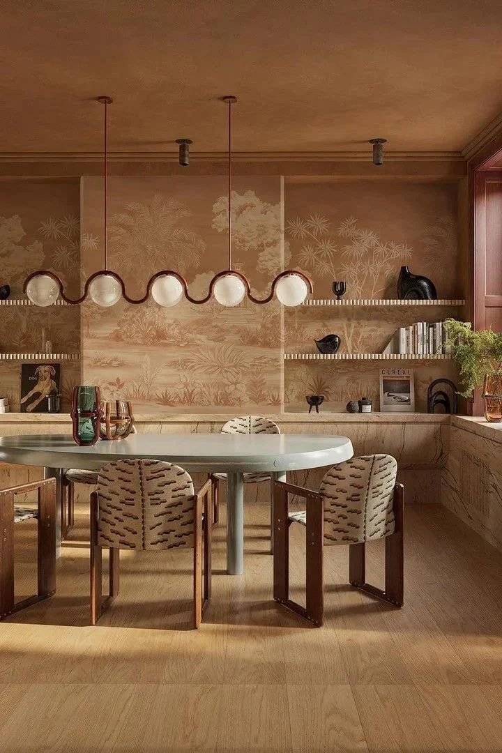 A dining room with a round table, four chairs with patterned upholstery, a mid-century modern chandelier, wooden shelves with decorative objects, books, and a window with a plants on the sill.