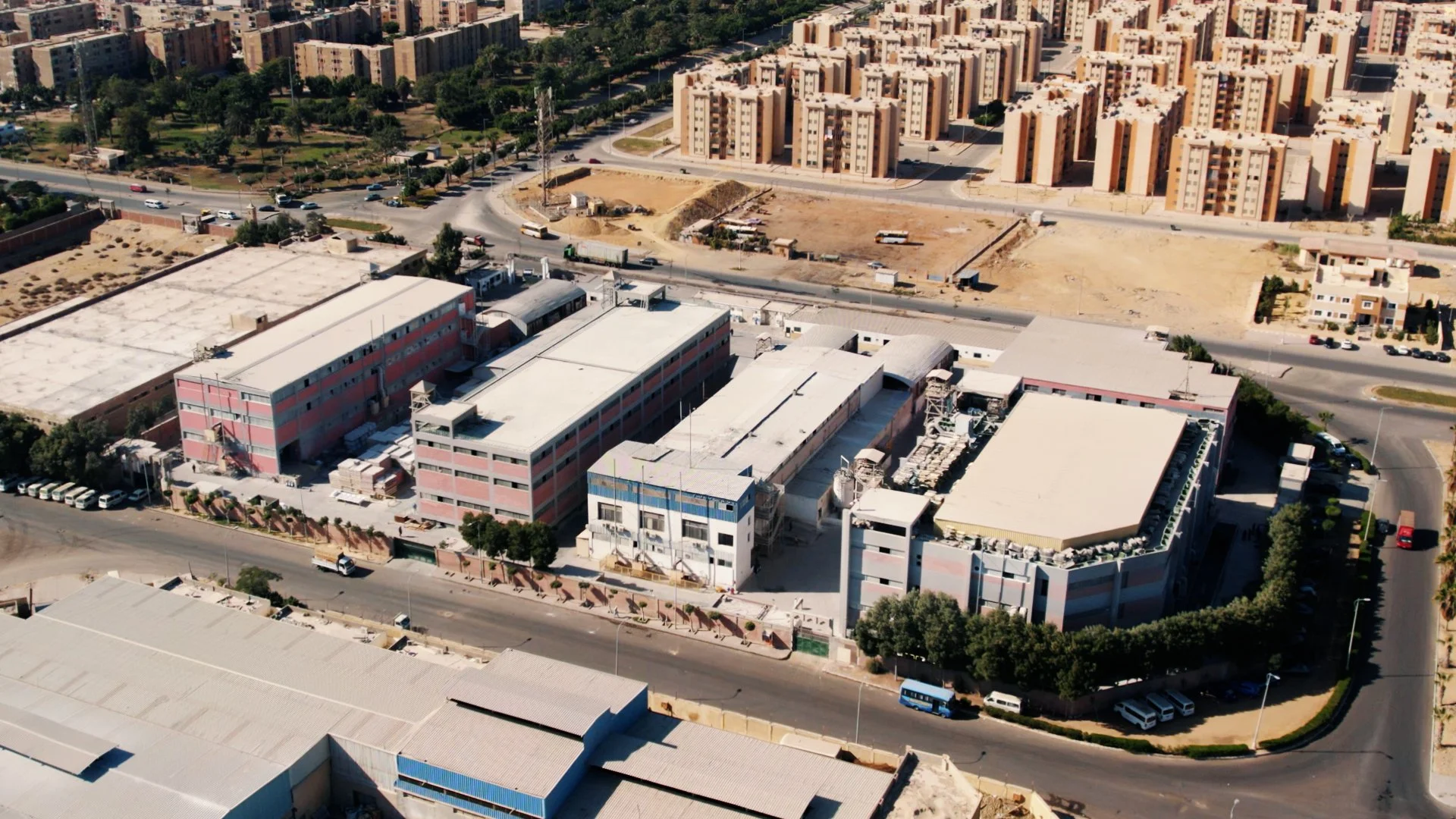 Aerial view of an urban area showing a large industrial or commercial complex with multiple buildings surrounded by roads, with residential buildings and open land in the background.