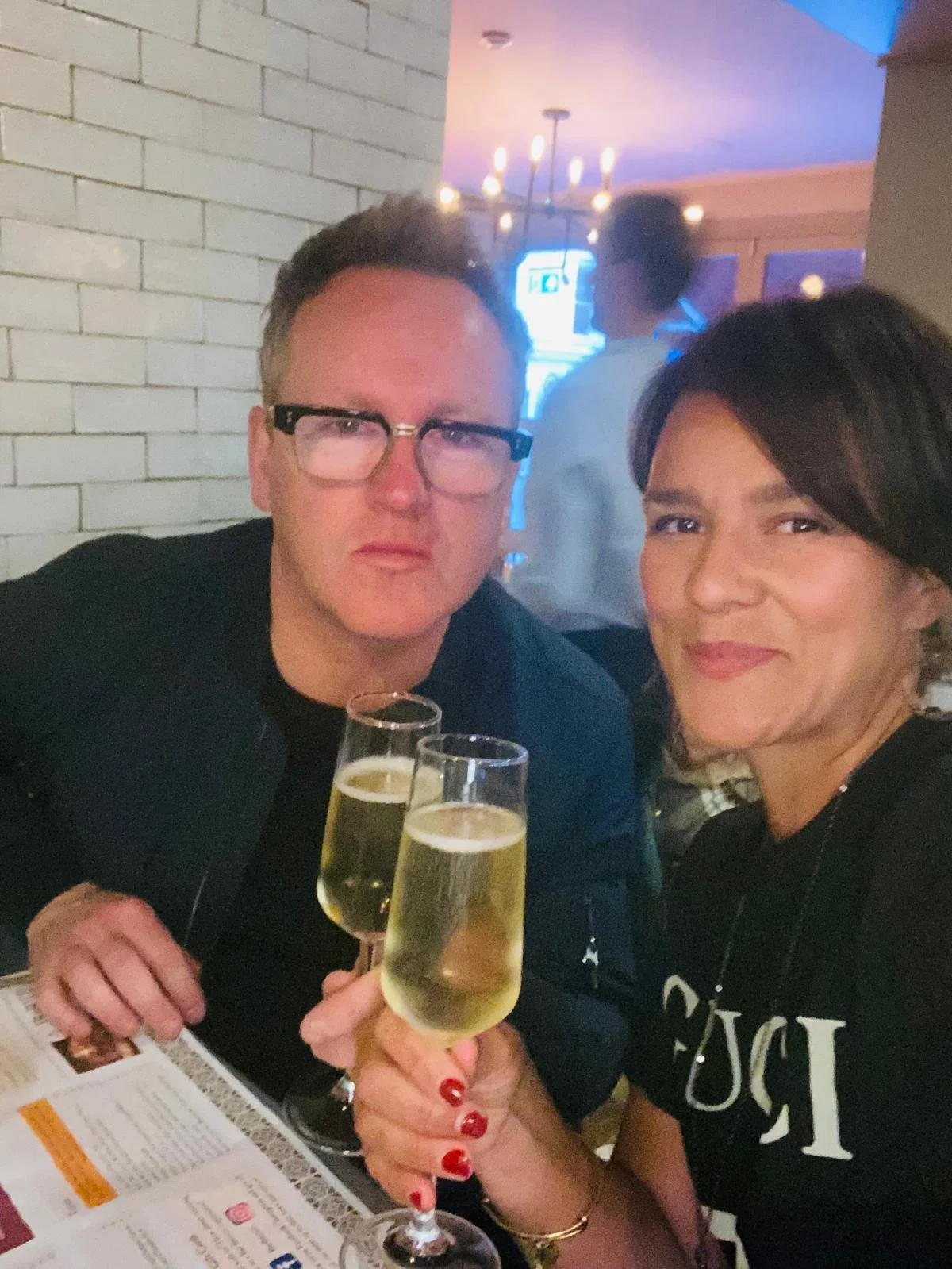 Two women and a man inside a restaurant or bar, holding glasses of champagne for a toast, with a white brick wall and a person in the background.
