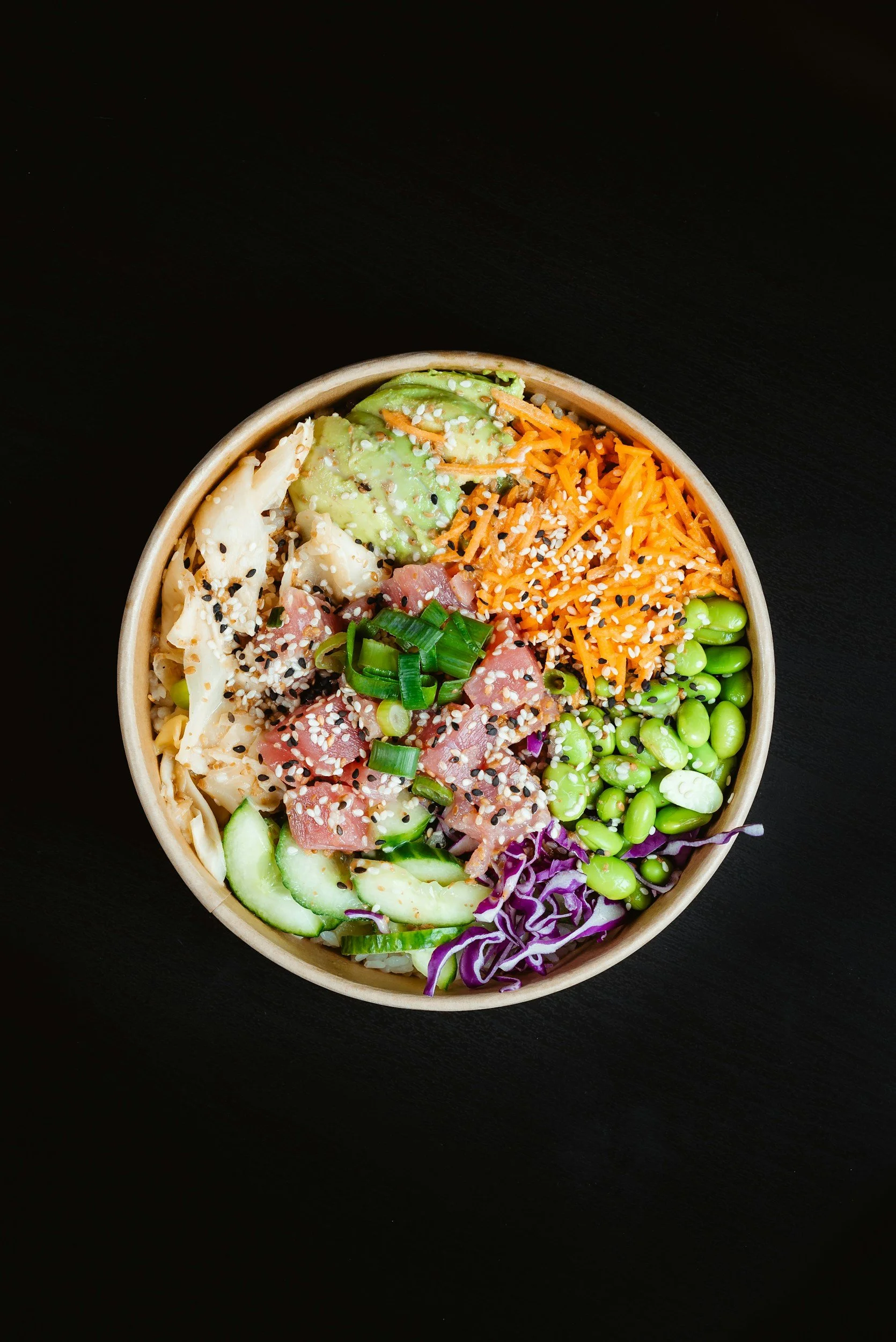 Bowl of mixed vegetables and sushi ingredients, including avocado slices, shredded carrots, edamame, cucumber slices, purple cabbage, chopped green onions, chopped raw fish. by Soul bowl