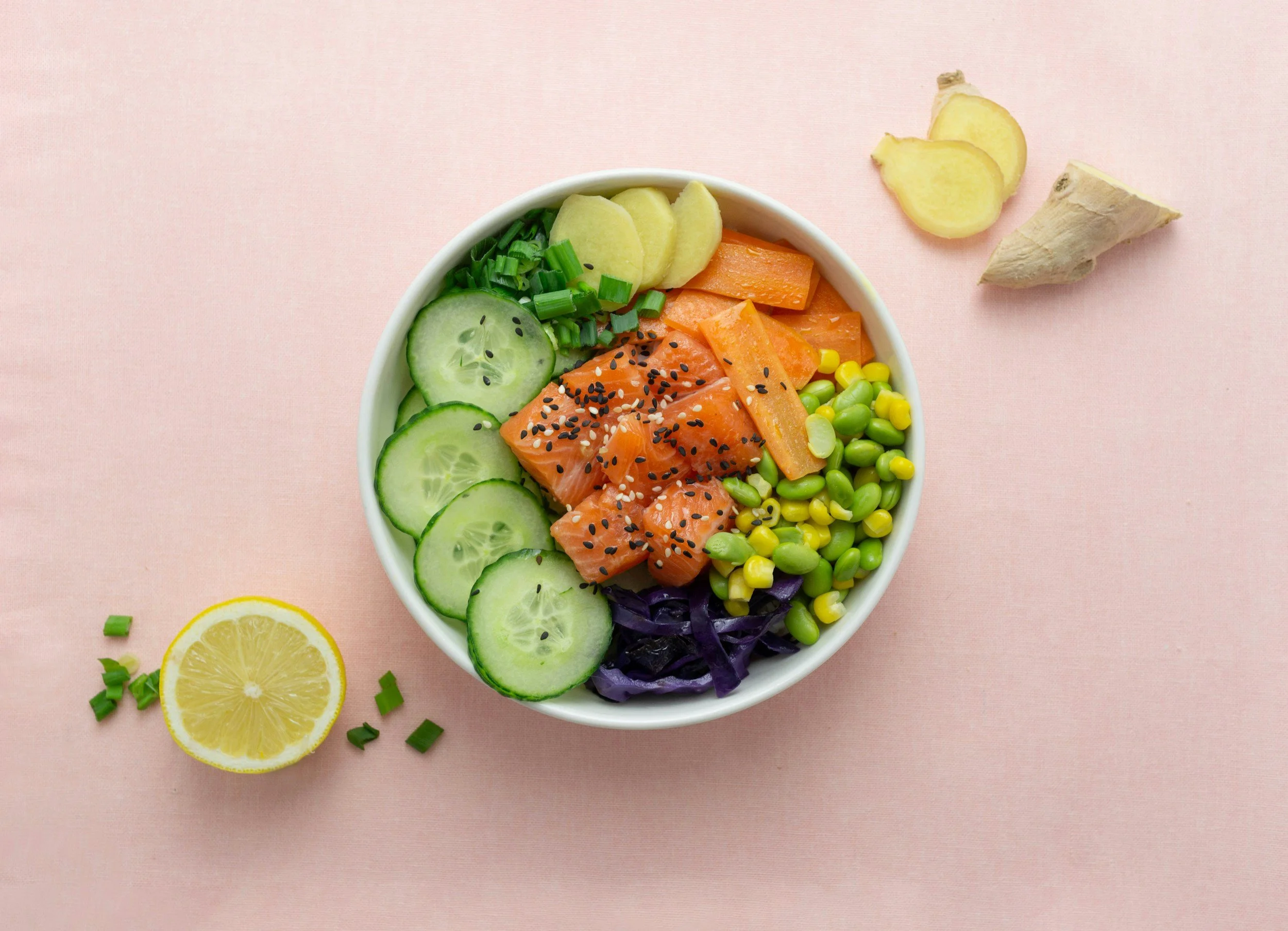 A bowl of colorful sushi ingredients including sliced cucumbers, green onions, ginger, orange salmon with black sesame seeds, yellow corn, and purple cabbage on a pink background from soul bowl