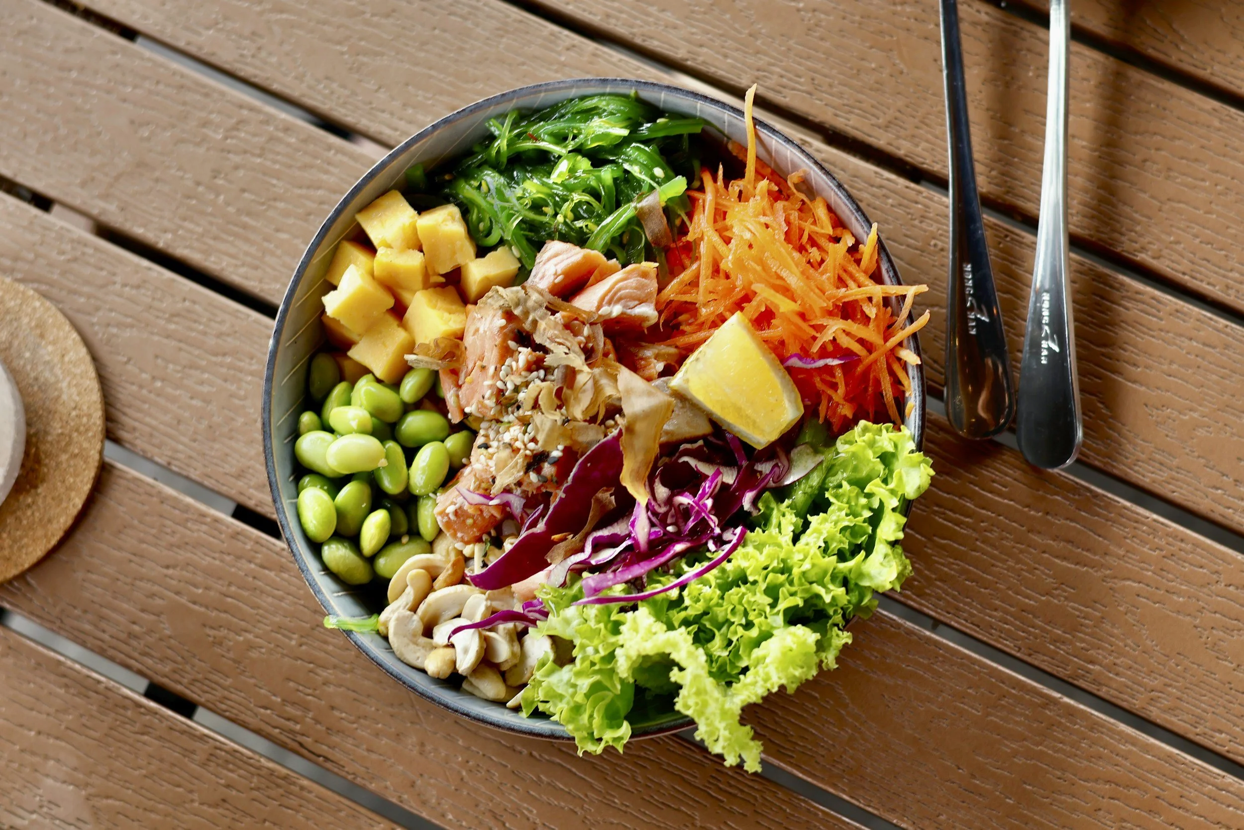 A bowl of Korean-inspired salad with diced potatoes, seaweed, shredded carrots, edamame, mixed greens, and salmon, placed on a wooden table with a pair of metal chopsticks. soul bowl poke bowls
