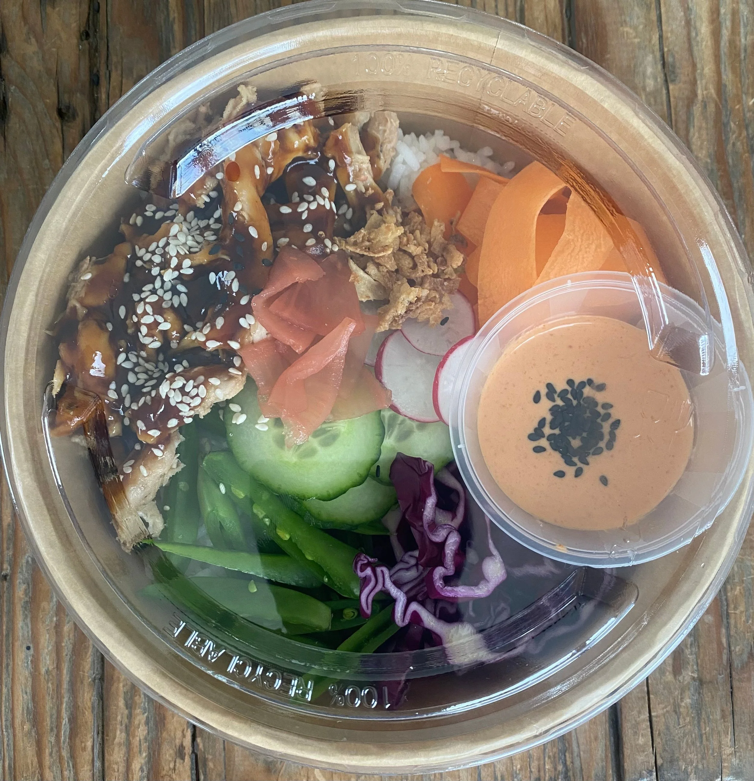A clear plastic takeout container with white rice, sliced cucumber, shredded purple cabbage, sliced radish, chopped pickled ginger, shredded lettuce, crispy wonton strips, teriyaki chicken with sesame seeds and sauce, and a small cup of pink sauce wi