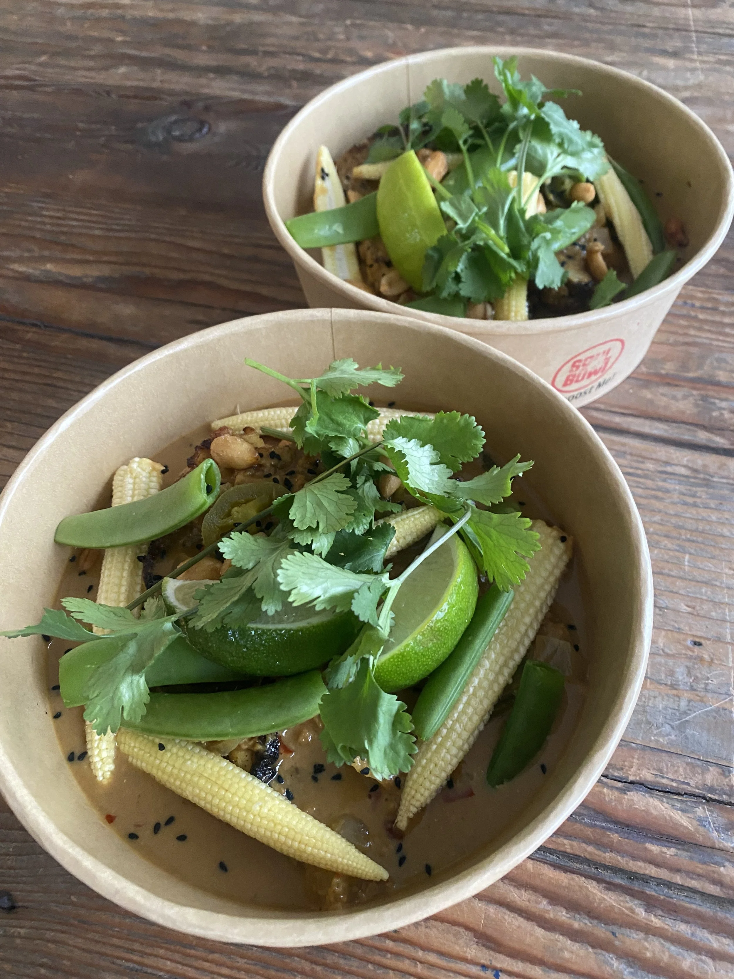 Two bowls of Asian-style vegetable soup topped with cilantro, lime wedge, snow peas, baby corn, and black sesame seeds, on a wooden table.