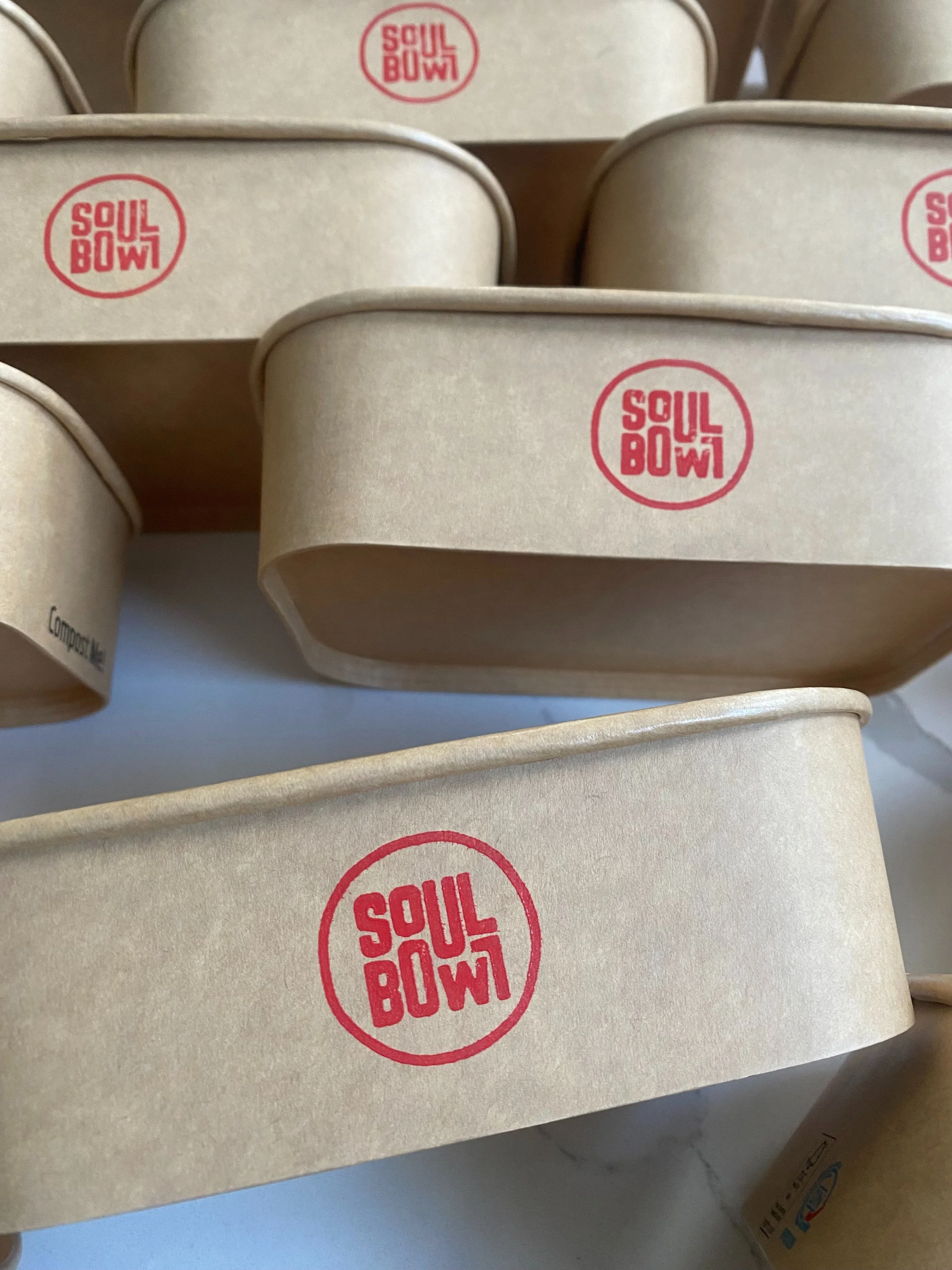 Multiple beige takeout containers with red 'Soul Bowl' logos stacked on top of each other. Soul Bowl take out, meal kit packaging