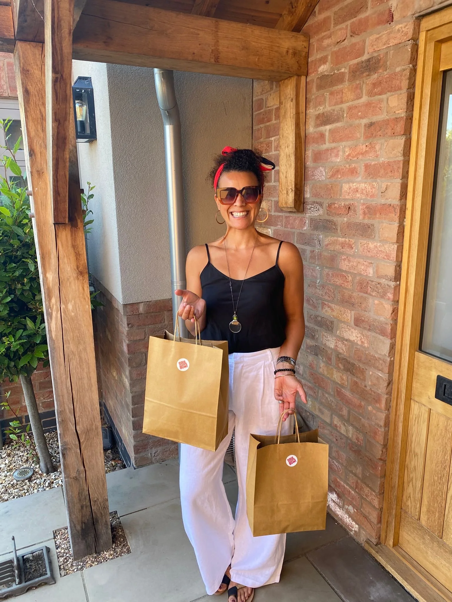 Smiling woman with curly hair wearing sunglasses, a red headband, a black sleeveless top, and white wide-leg pants, holding two brown paper shopping bags, standing outside near brick and wooden walls.