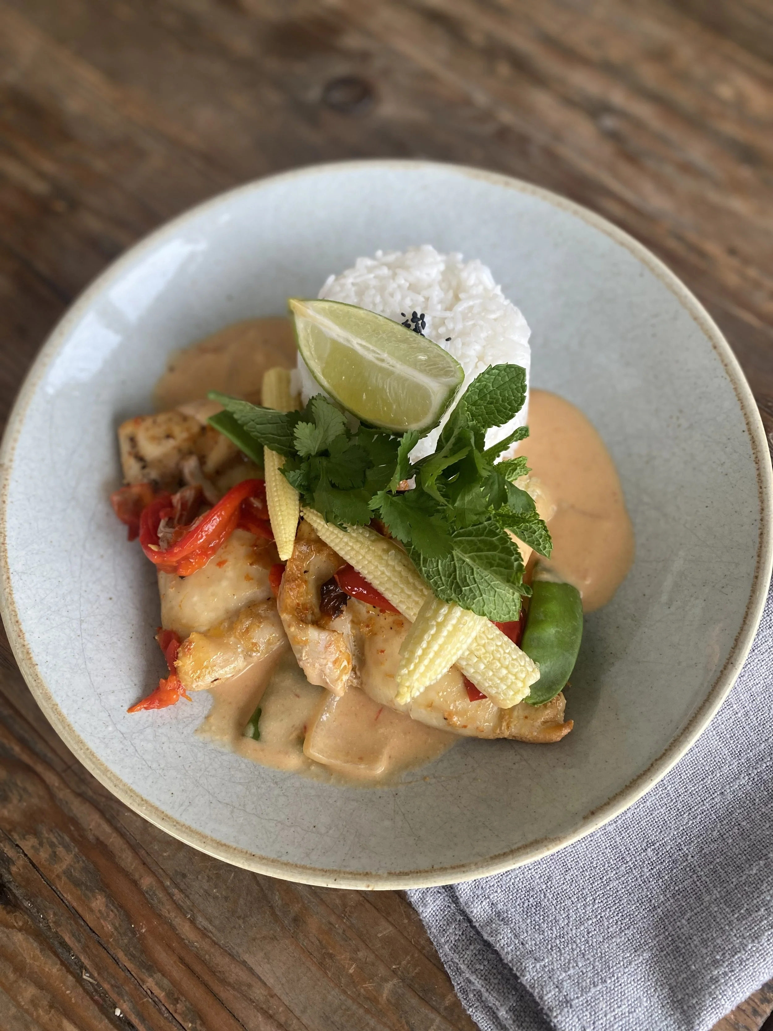 A bowl of Thai green curry with rice, garnished with a lime wedge, fresh herbs, and vegetables.