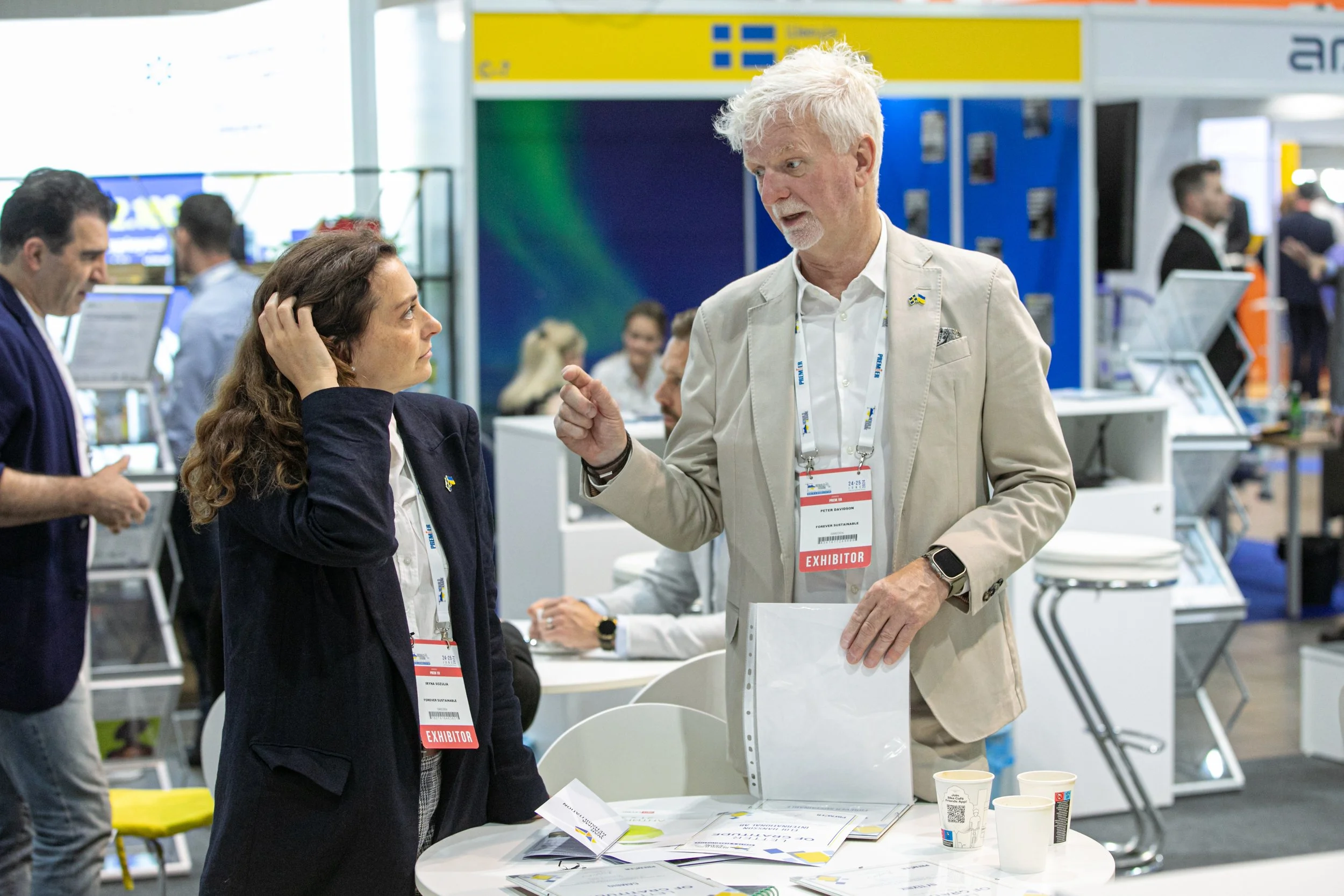 Two people, a woman and a man, talking at an exhibition booth. The woman has brown hair, and the man has white hair and a beard. Both are wearing conference badges that say 'Exhibitor'. The man appears to be explaining something, and the woman is listening attentively. There are brochures, paper cups, and a clear plastic folder on the table in front of them, with other attendees and booths visible in the background.