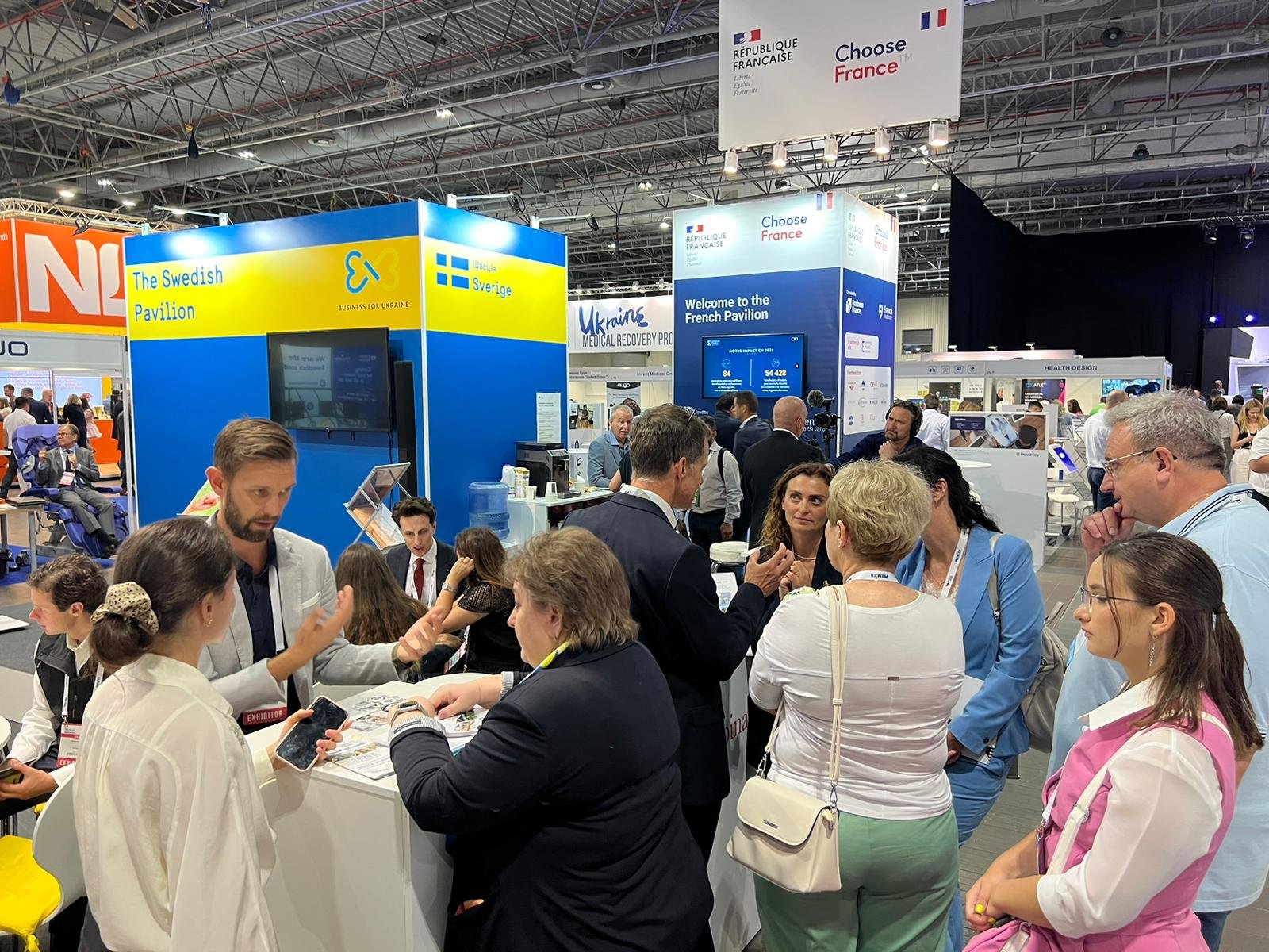 Various people attending and engaging at an international exhibition booth labeled 'The Swedish Pavilion' with flags and signage indicating countries like France, Sweden, and Ukraine, and various informational displays in the background.
