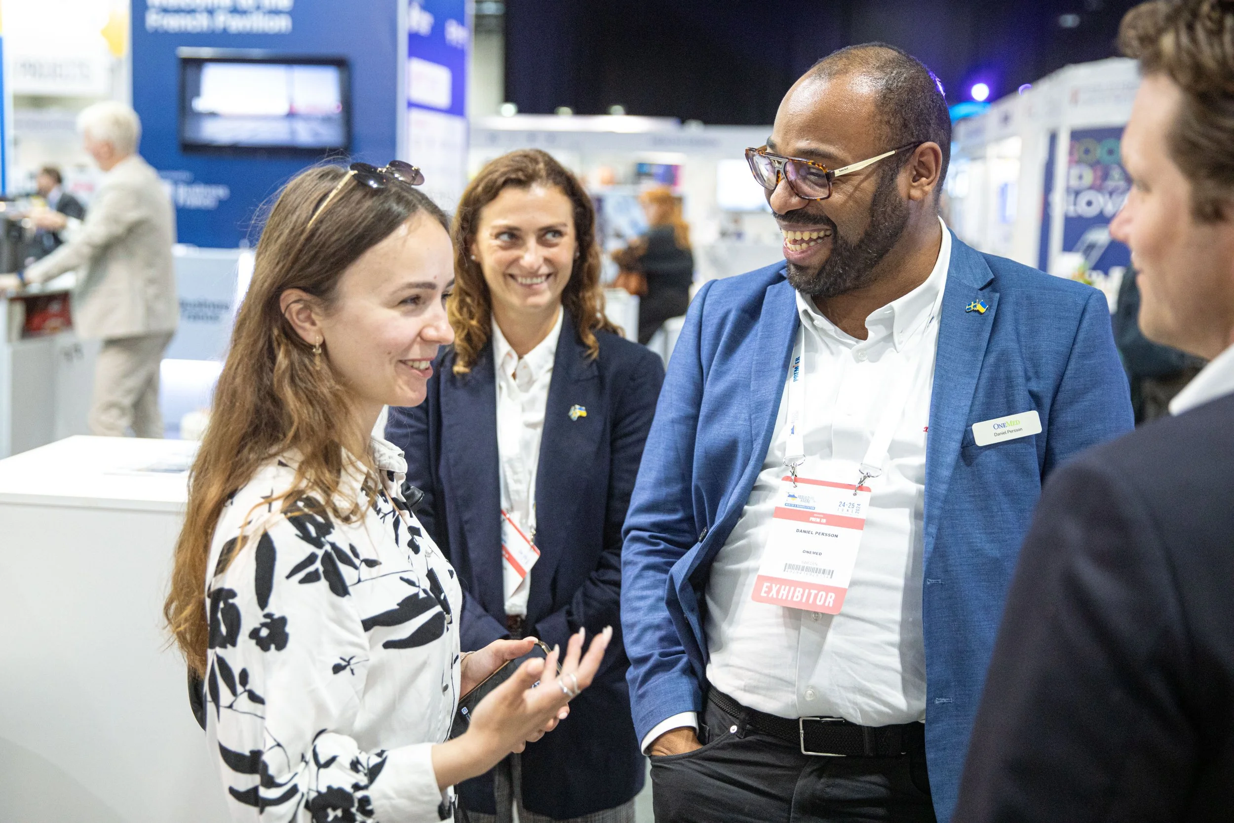 Four people smiling and conversing at a professional event or conference. The woman on the left is holding a phone, and the man on the right is wearing a badge labeled 'EXHIBITOR'. The background shows exhibition booths and a monitor.