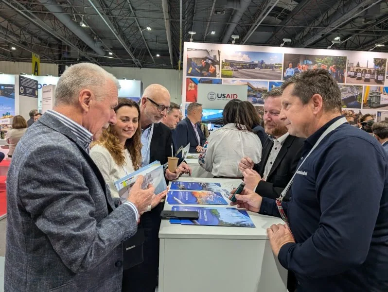 Group of people at a trade show or conference, engaging with brochures and displays. US AID logo visible in background.