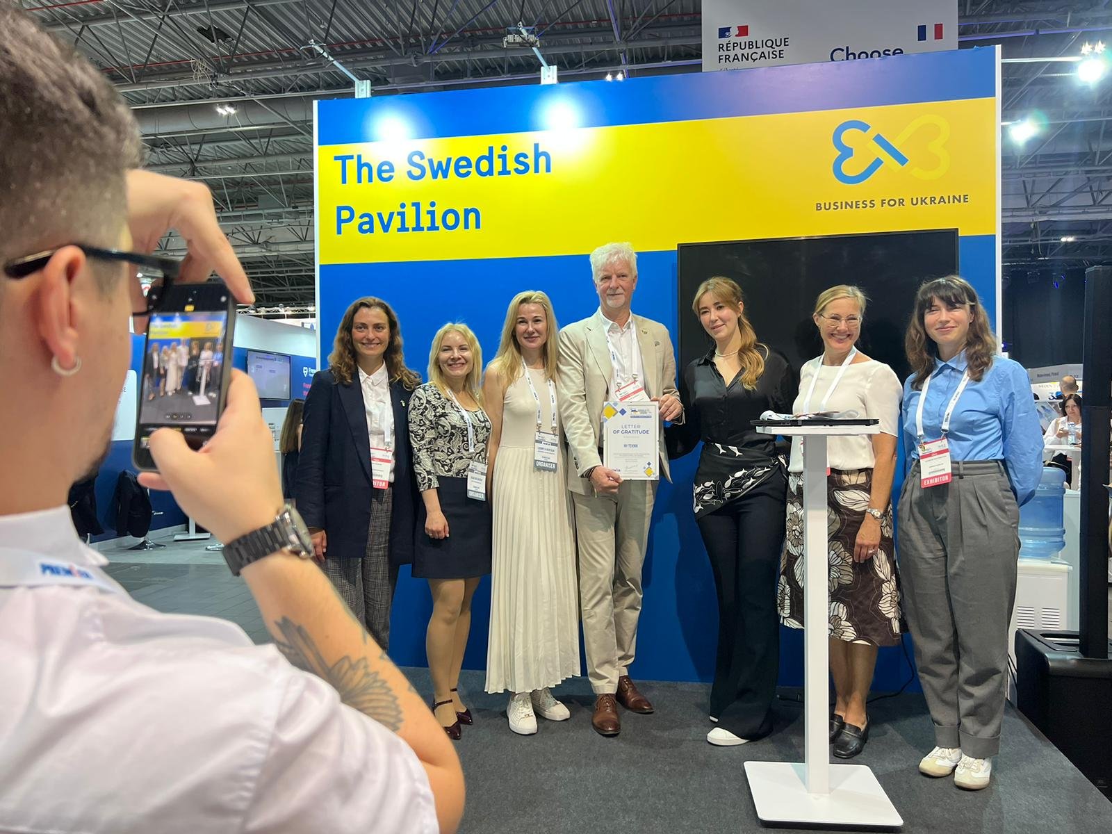 Group of eight people standing in front of for a photo at a Swedish Pavilion booth, with one person taking a picture, at an indoor event. The background has a blue and yellow sign that reads 'The Swedish Pavilion' and a logo with the words 'Business for Ukraine.' The group is smiling, with some people holding papers or certificates.