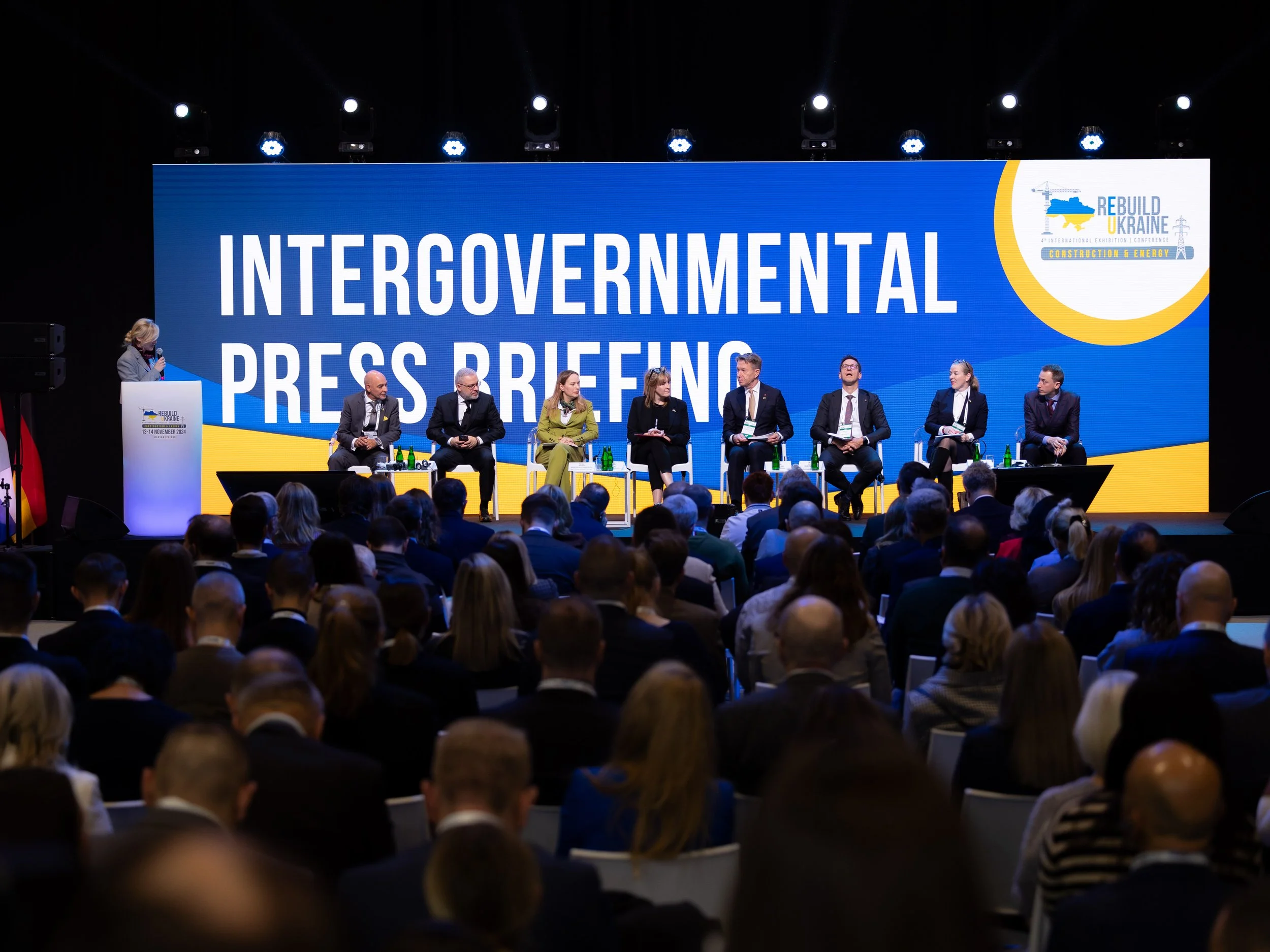 A large conference stage with a panel of seven people seated and a woman speaking at a podium. Behind them is a big screen displaying 'Intergovernmental Press Conference' and a logo for 'Rebuild Ukraine' and 'Construction & Energy.' The audience is seated in front, facing the stage.