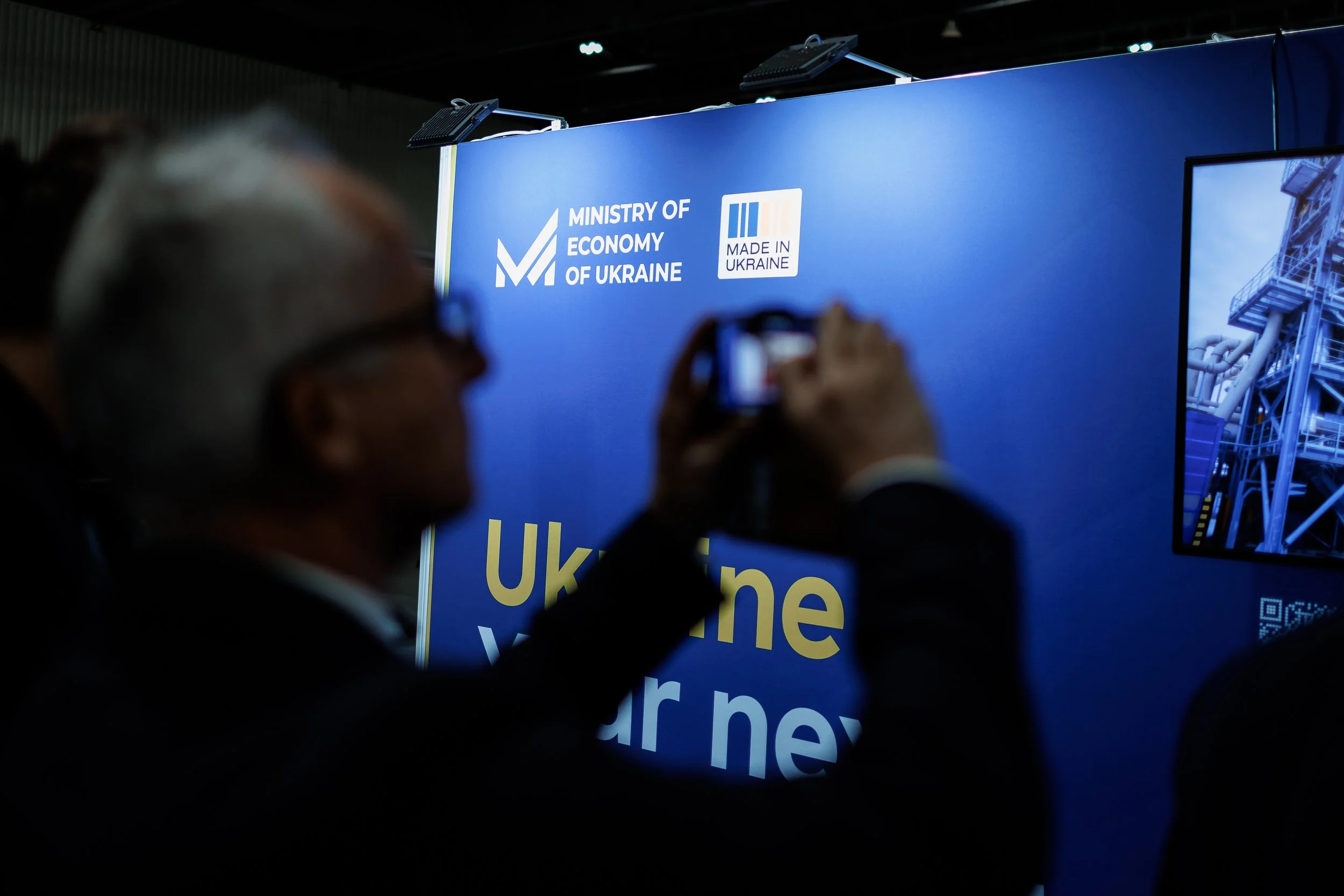 Person taking a photo of a blue display board with logos of the Ministry of Economy of Ukraine and Made in Ukraine, at an event.
