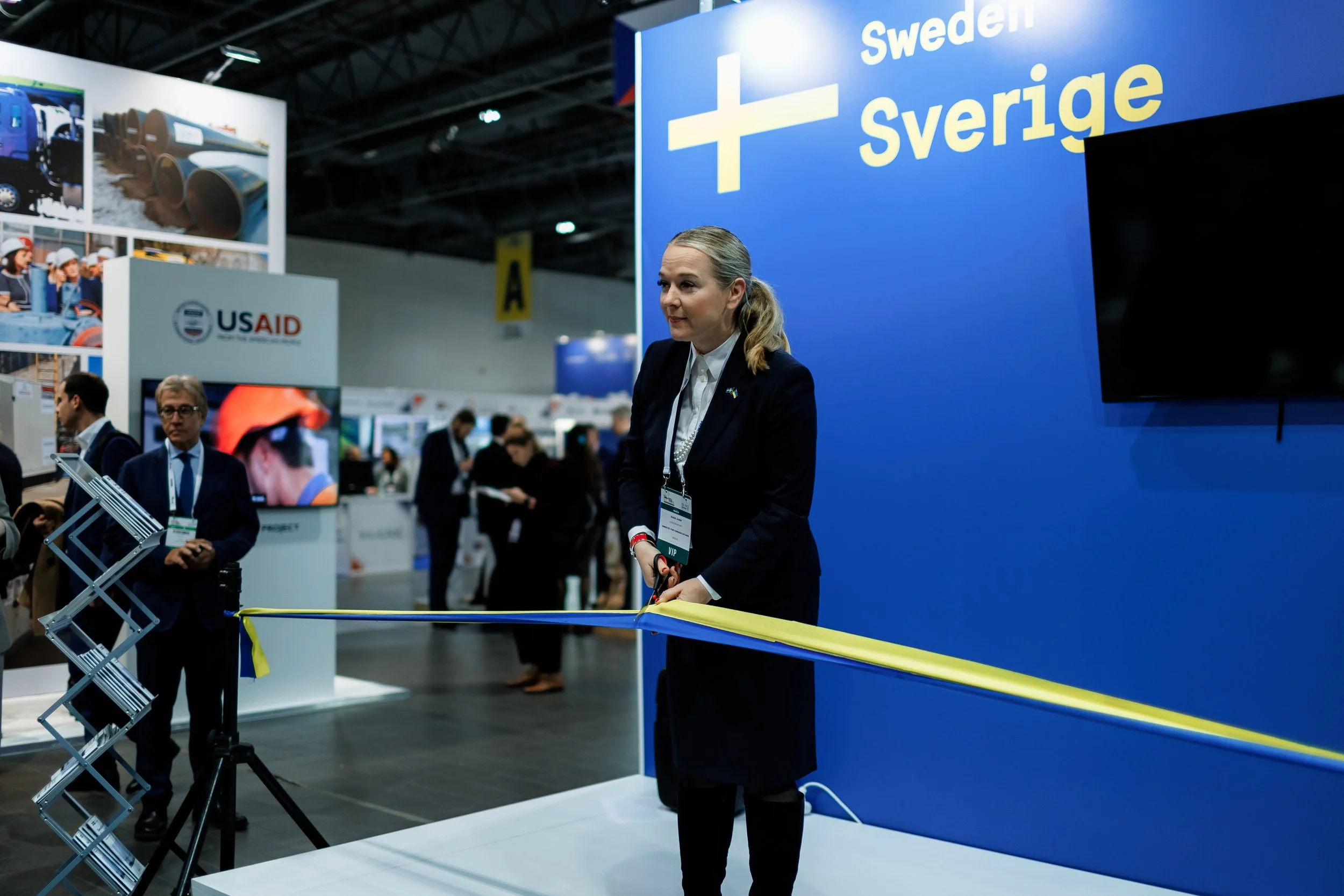 A woman in a black suit cutting a ribbon with scissors at an event booth marked 'Sweden' or 'Sverige,' with multiple people in the background.