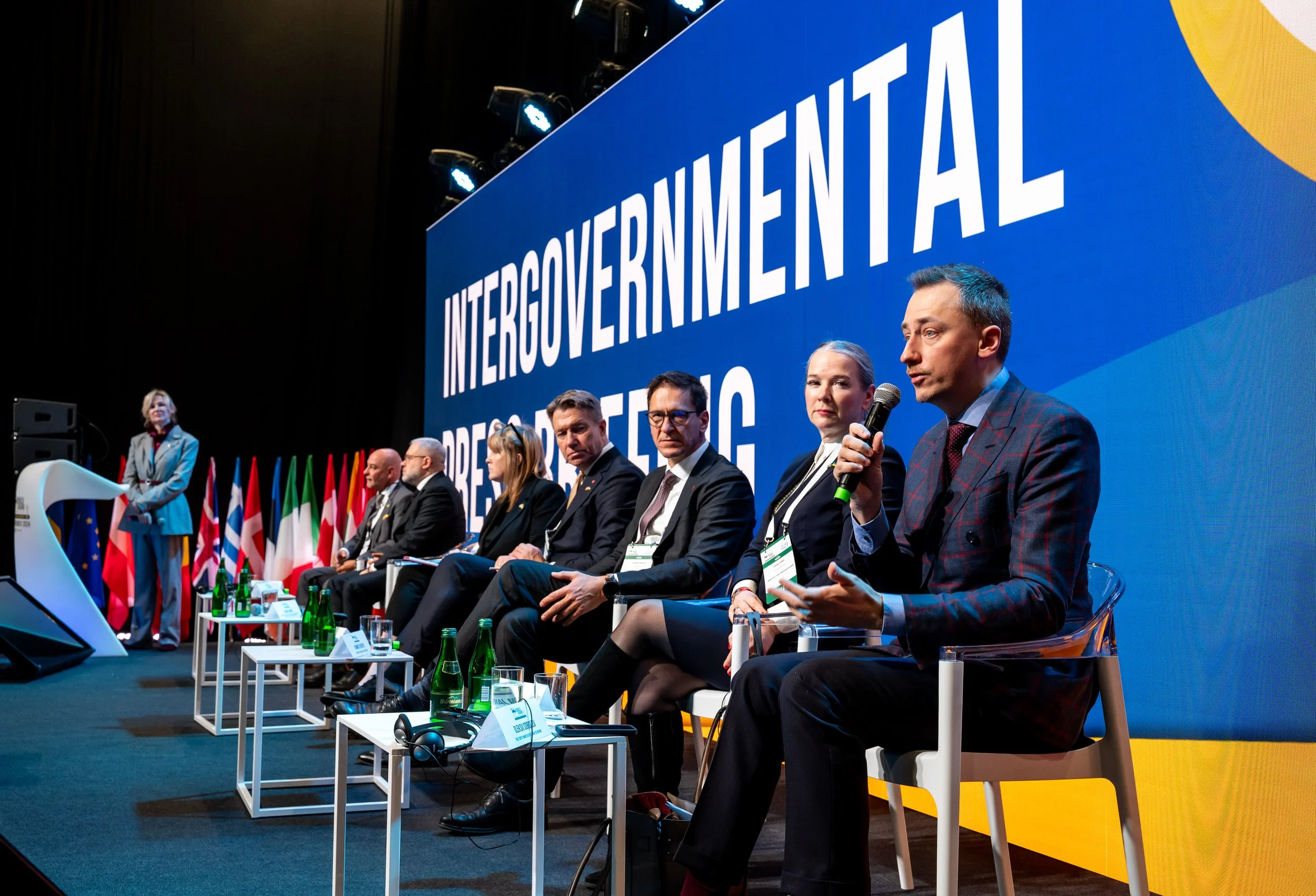 Panel of seven people sitting on stage with a large screen behind them displaying the words 'INTERGOVERNMENTAL' and 'REGULATORY.' A woman is standing at a podium on the left. Several flags are displayed along the stage. The speaker on the right is ho