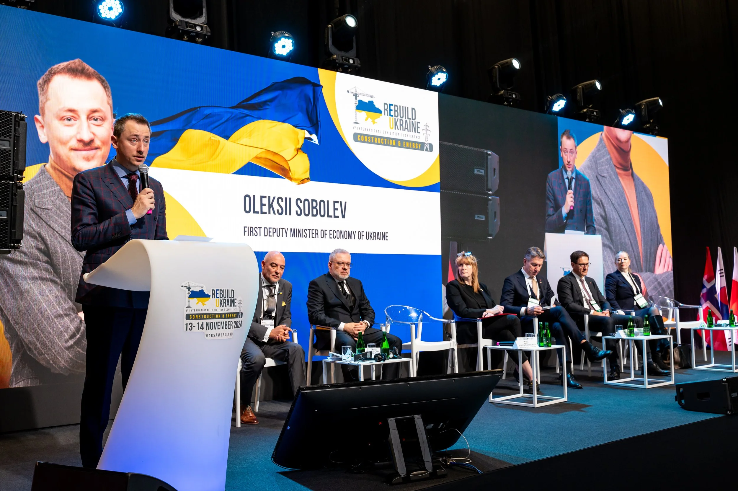 A man speaking at a podium on stage during a conference about rebuilding Ukraine, with a large screen behind him displaying his name, Oleksii Sobolev, First Deputy Minister of Economy of Ukraine. Several people are seated on stage, and the screen sho