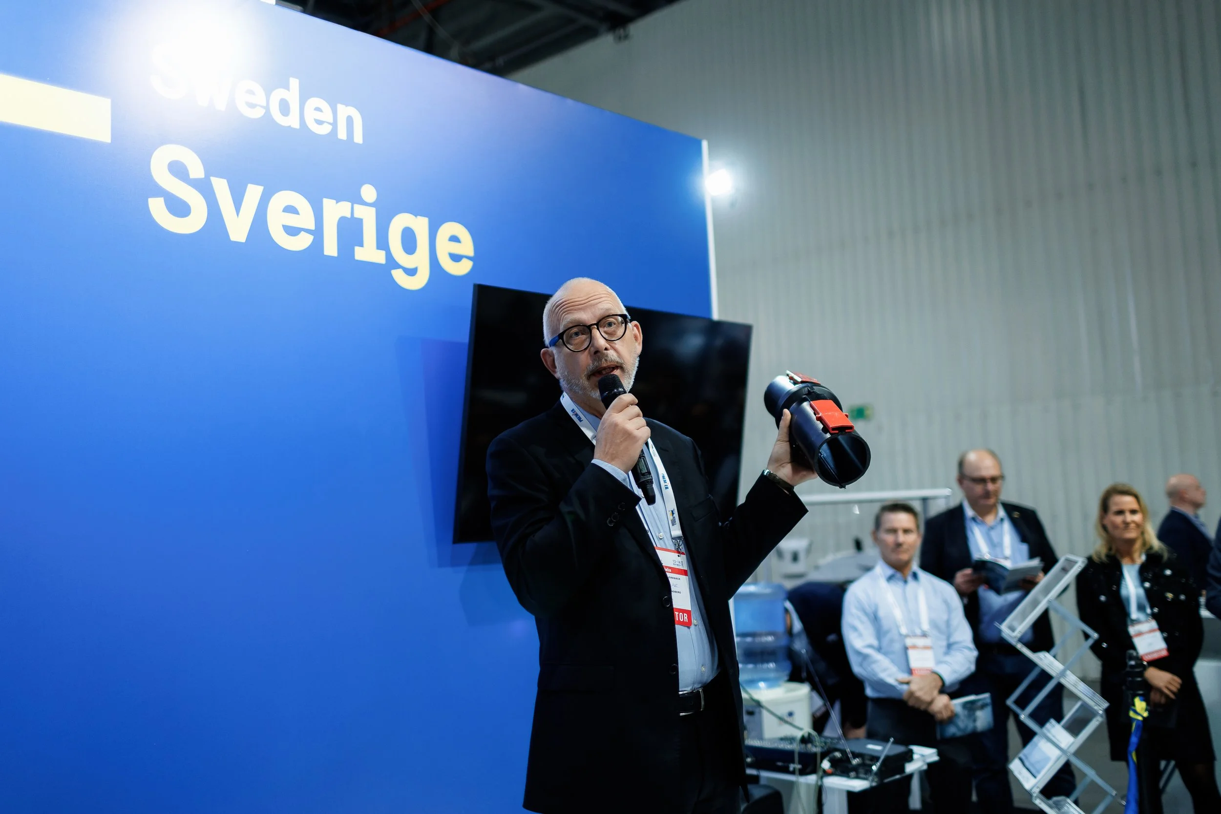 A man in a suit holding a microphone and a telescope at an international trade exhibit, with a blue backdrop that reads 'Sweden' and 'Sverige', and several people in the background.