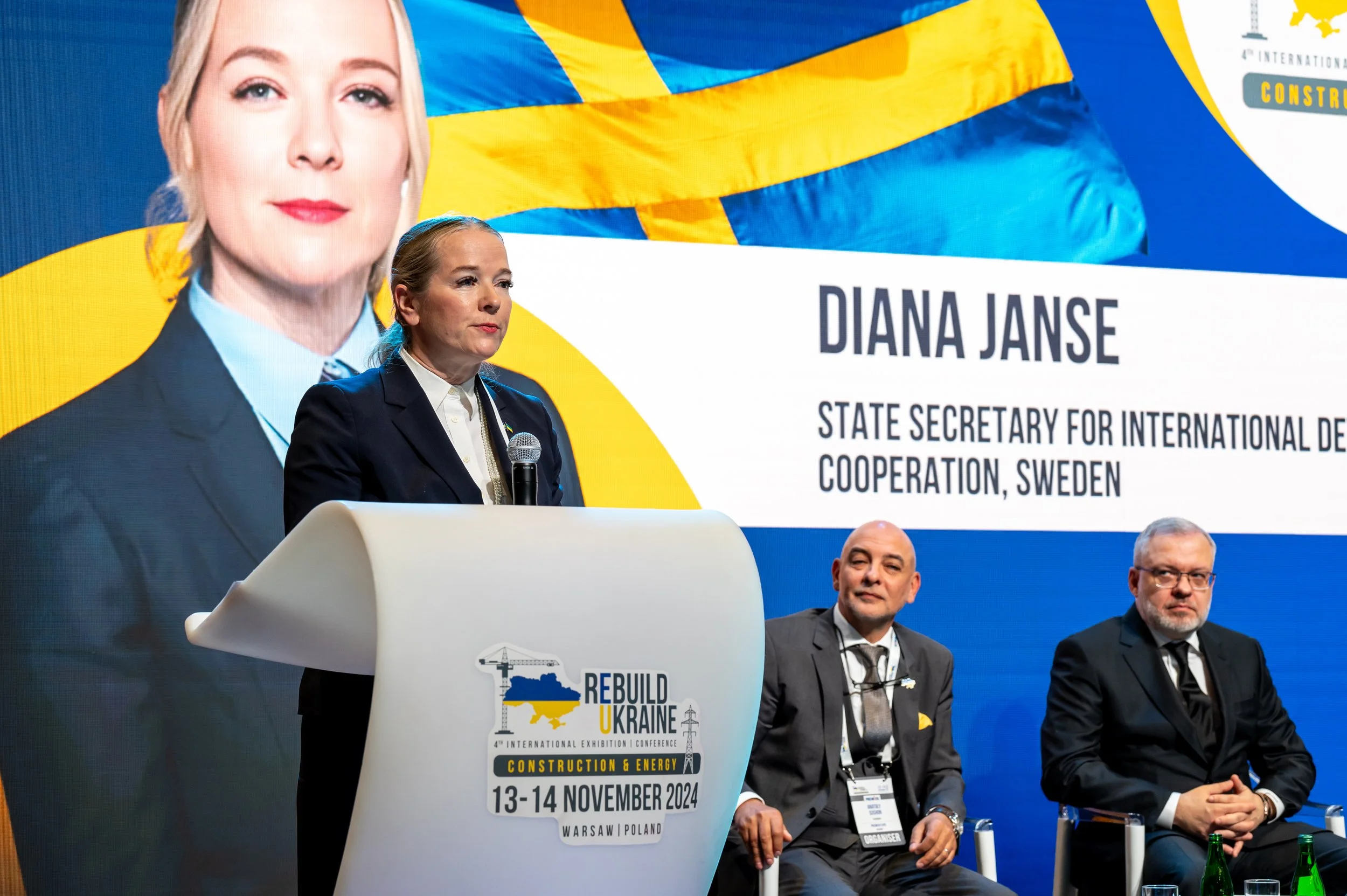 Diana Janse speaking at a conference podium with a large screen behind her displaying her name and title. Two men are seated beside her on stage.