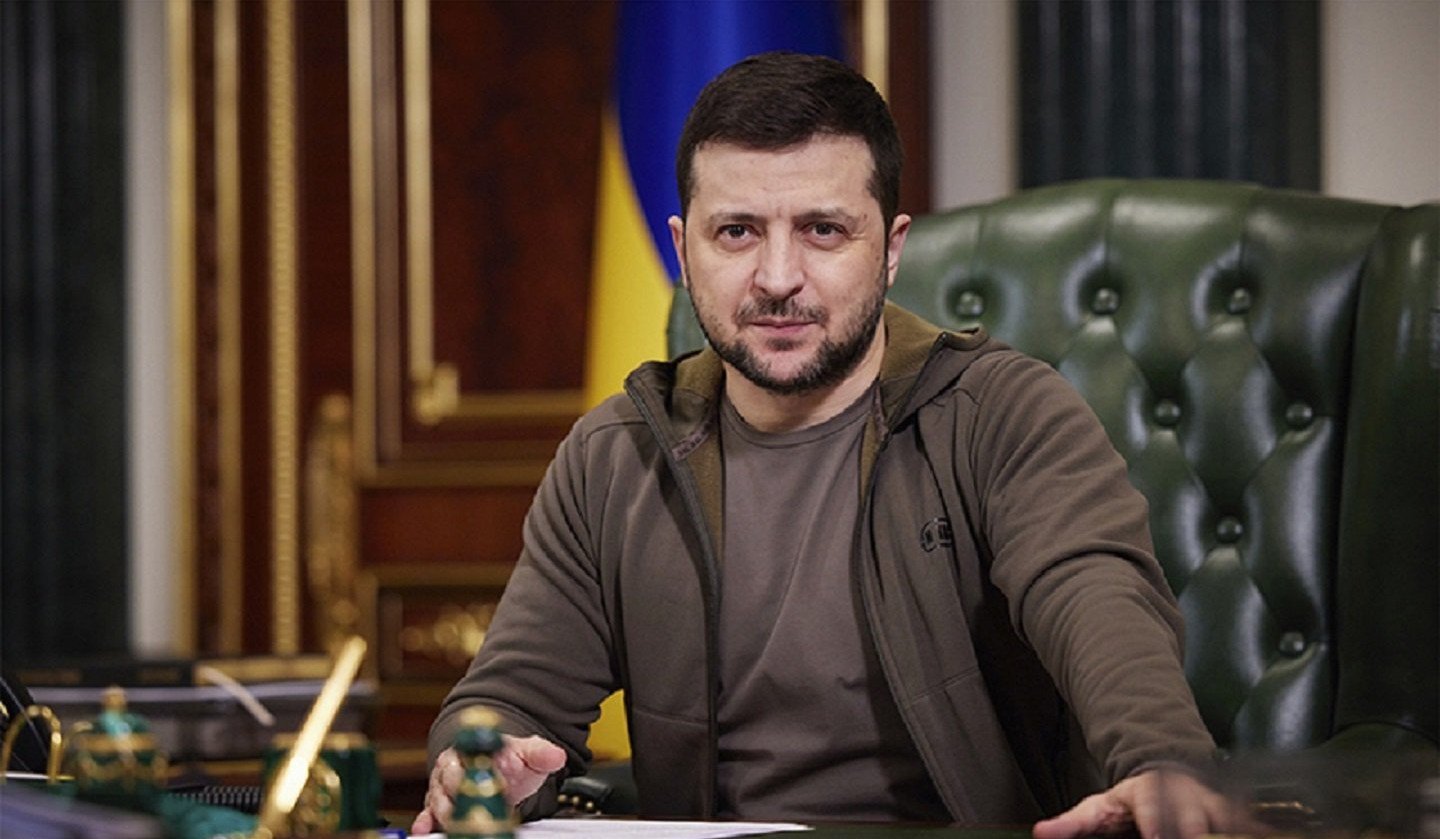 The president of Ukraine sitting at a desk with a Ukrainian flag in the background, wearing a brown jacket.