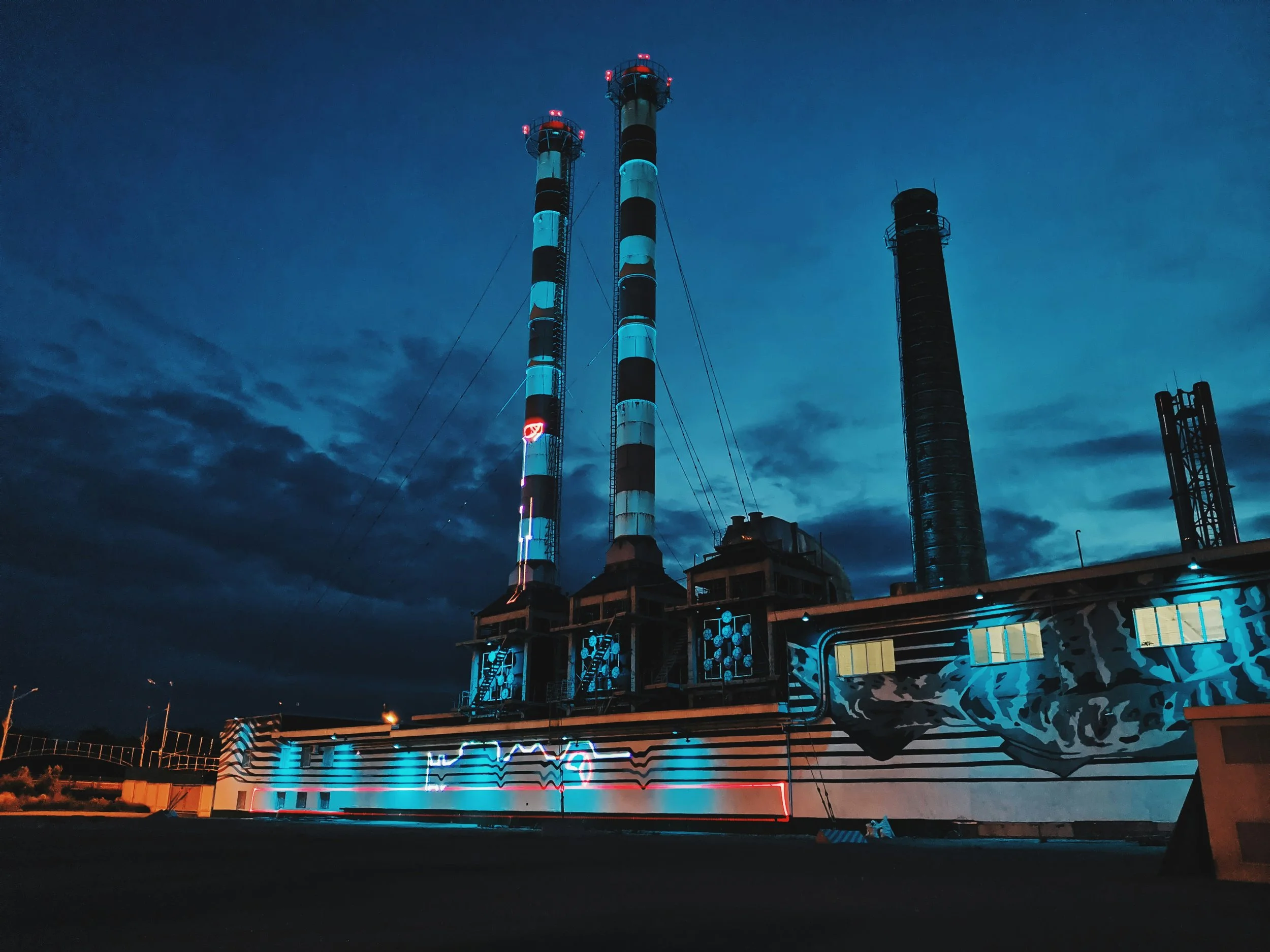 Nighttime view of an industrial facility with three tall smokestacks, illuminated with blue neon lights and artwork on the building exterior, under a dark, cloudy sky.
