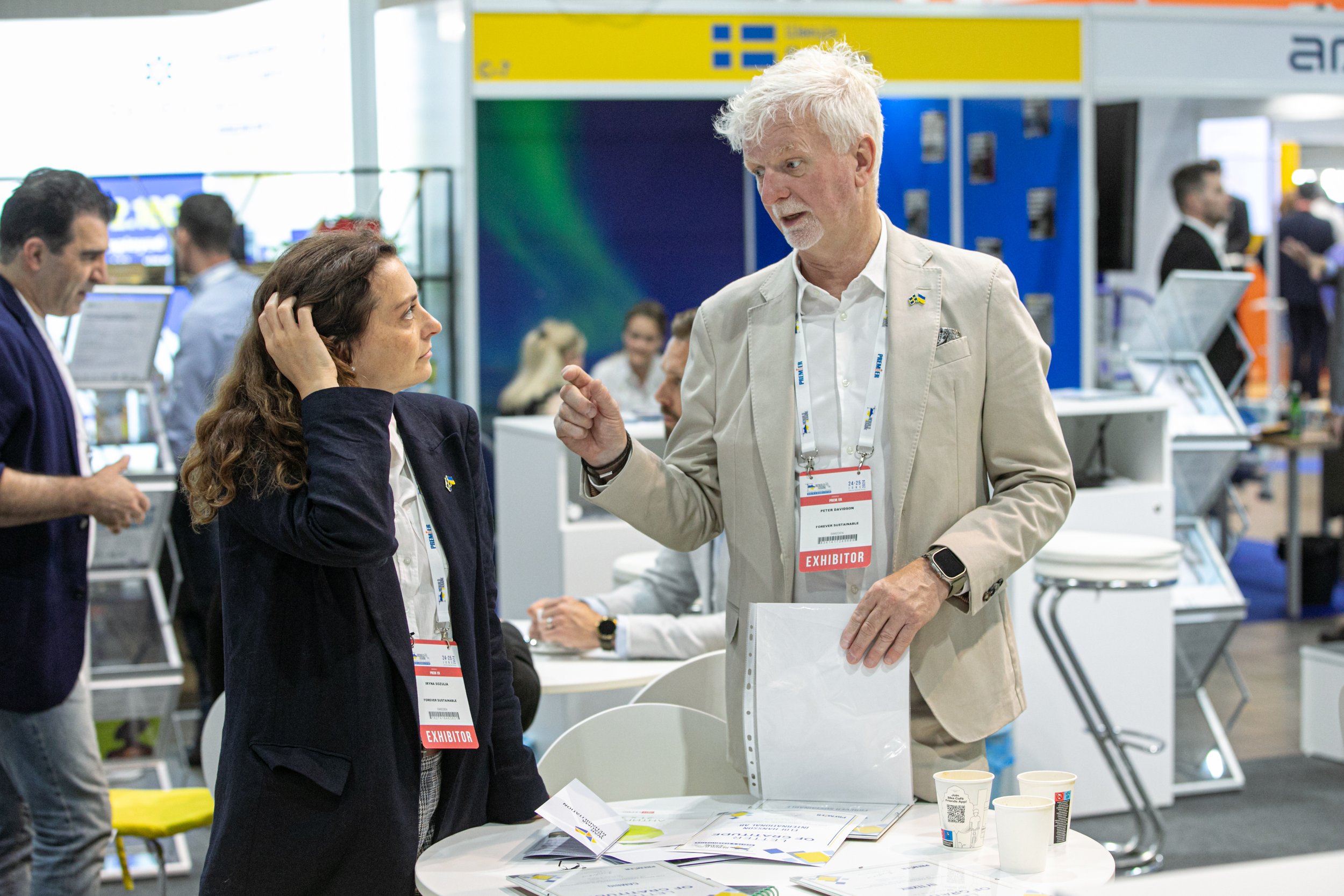 Two business professionals at a conference, engaged in conversation at a booth with exhibit badges, with several other attendees and booths visible in the background.