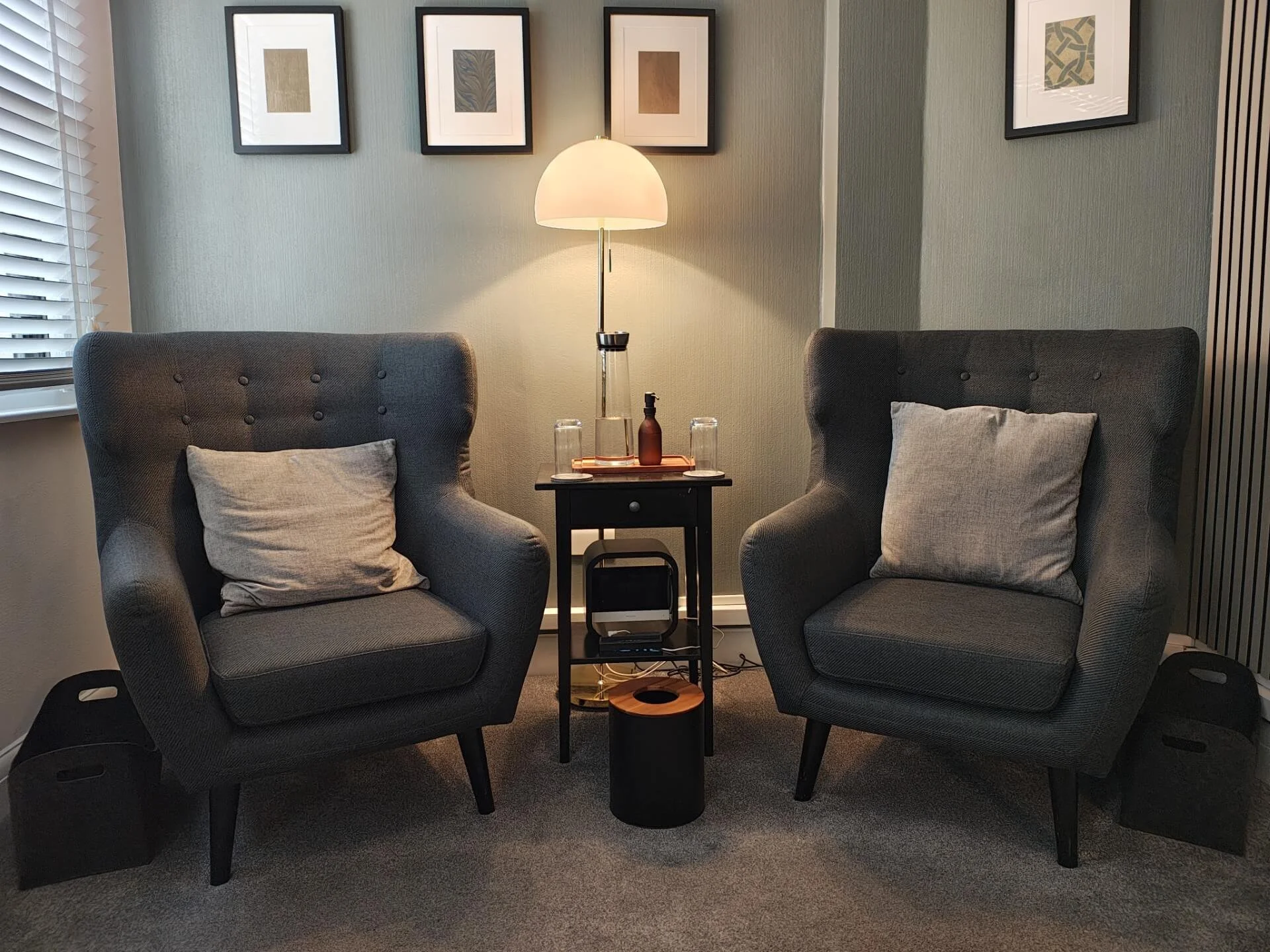 Two gray armchairs with beige pillows facing each other, a small black side table with a lamp, a brown glass bottle, and two glasses, and four framed abstract art pieces on the wall behind, in a cozy living room corner.