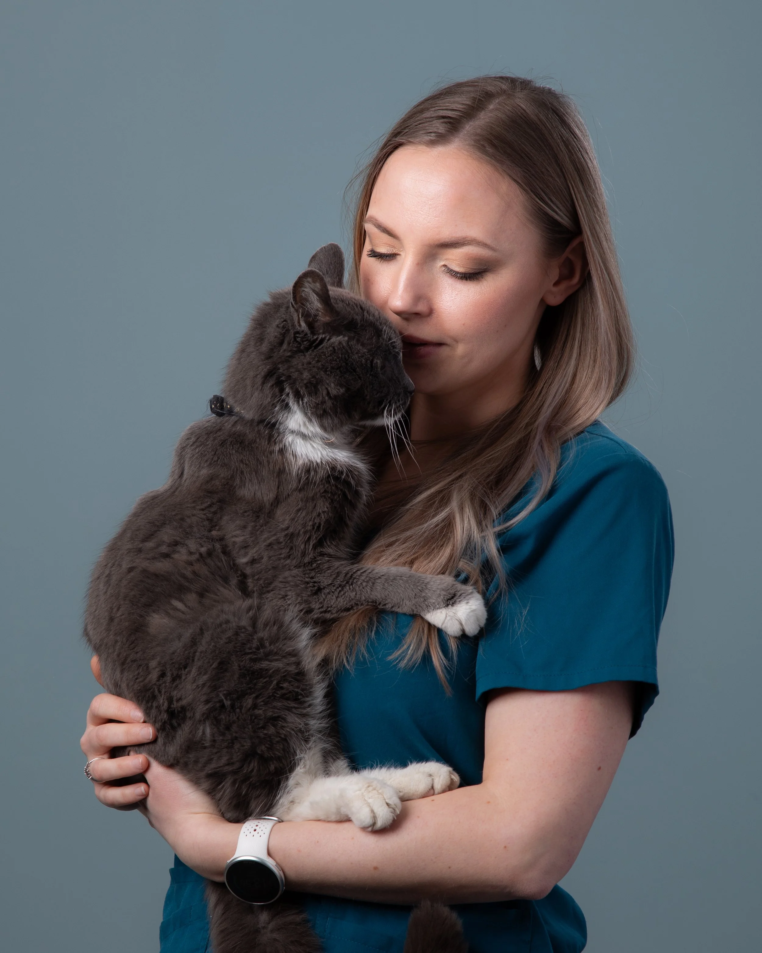 En ung kvinne som holder en stor, grå og hvit katt nært ansiktet og kysser den. Kvinnen har langt, lyst hår og bruker en blå sykepleieruniform.