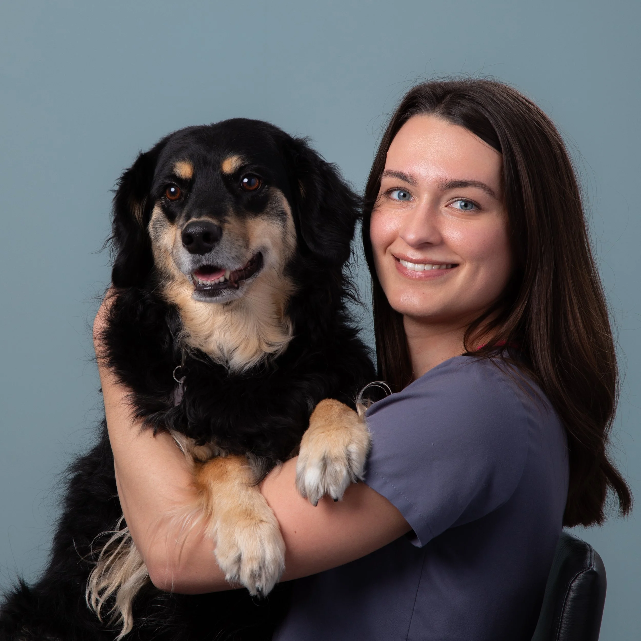 En kvinne holder en stor svart og brun hund med hvite detaljer. Begge ser glade ut mot en lys blå bakgrunn.