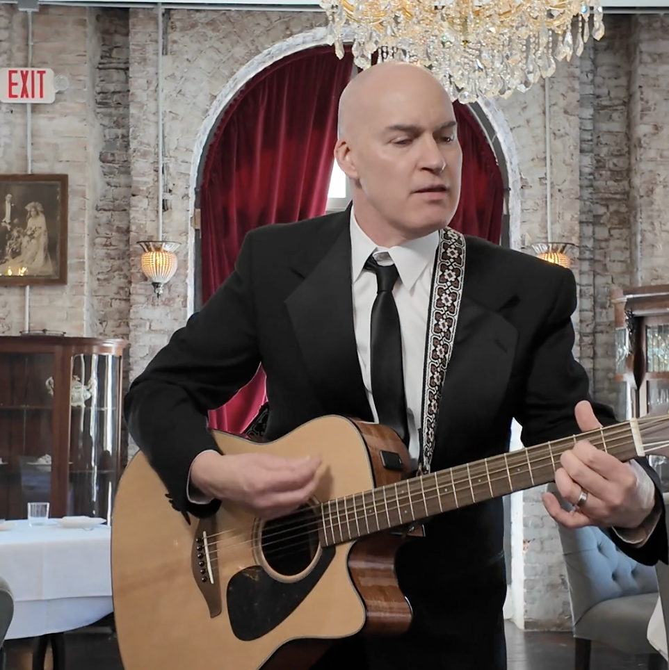 A bald man dressed in a black suit with a patterned guitar strap, playing an acoustic guitar in a restaurant or banquet hall with exposed brick walls, red curtains, and a chandelier.