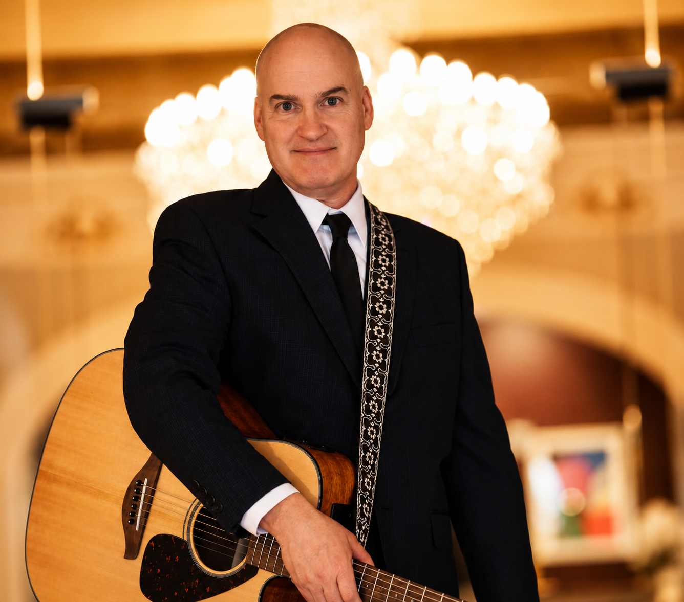 A man in a black suit with a white shirt and black tie holding an acoustic guitar, standing in a warmly-lit room with a chandelier in the background.